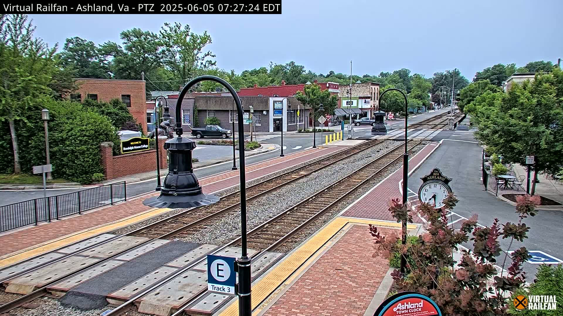 A mostly sunny view of a train track running through a town, with brick sidewalks, landscaping, and buildings lining the street.