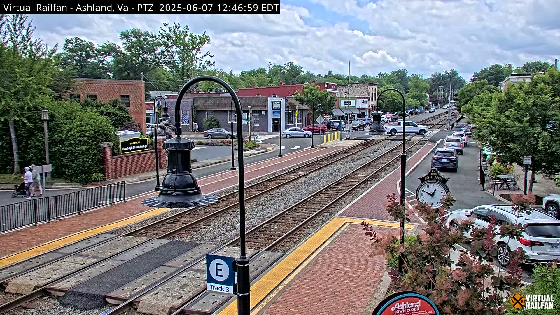 A partly cloudy day reveals a train track running through a town center, with cars parked along the street and people walking near a college campus.