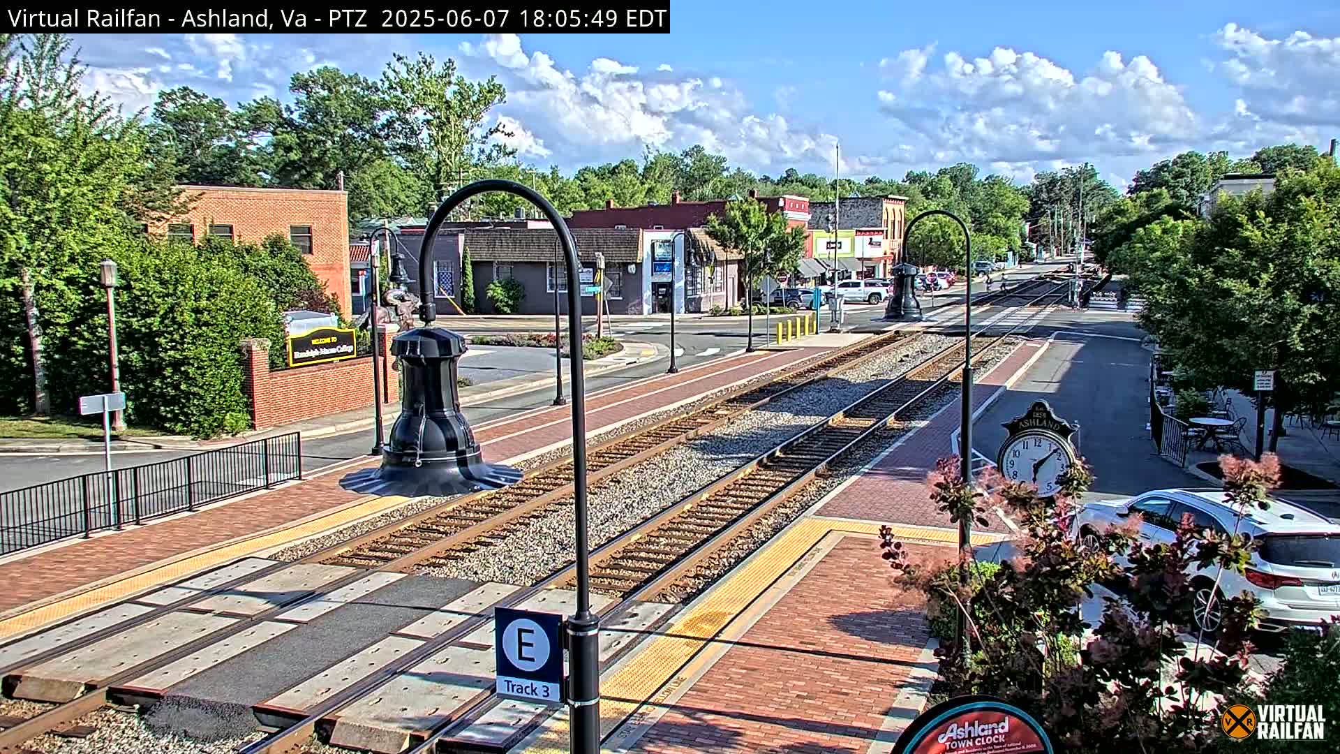 A mostly sunny day shows a train track running through a town, with brick sidewalks, buildings, and greenery lining the street.