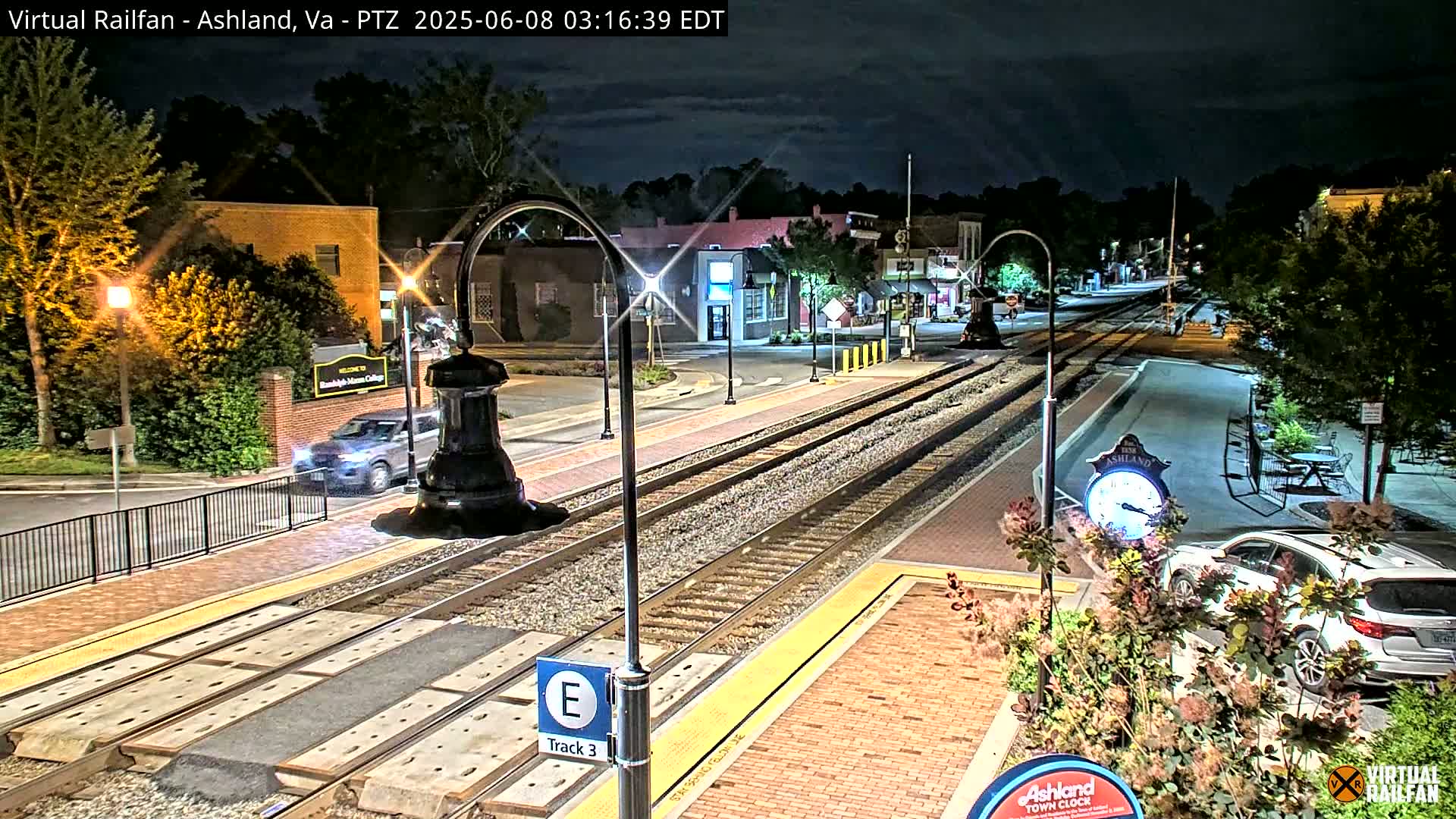A nighttime view of a train station platform and town square, with train tracks running through the center, under a partly cloudy sky.
