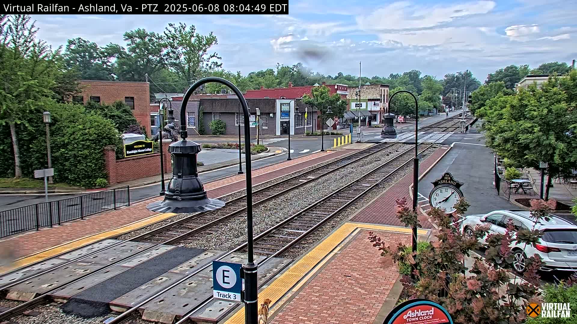A partly cloudy day reveals train tracks running through a town center, with brick sidewalks, lampposts, and buildings visible in the background.
