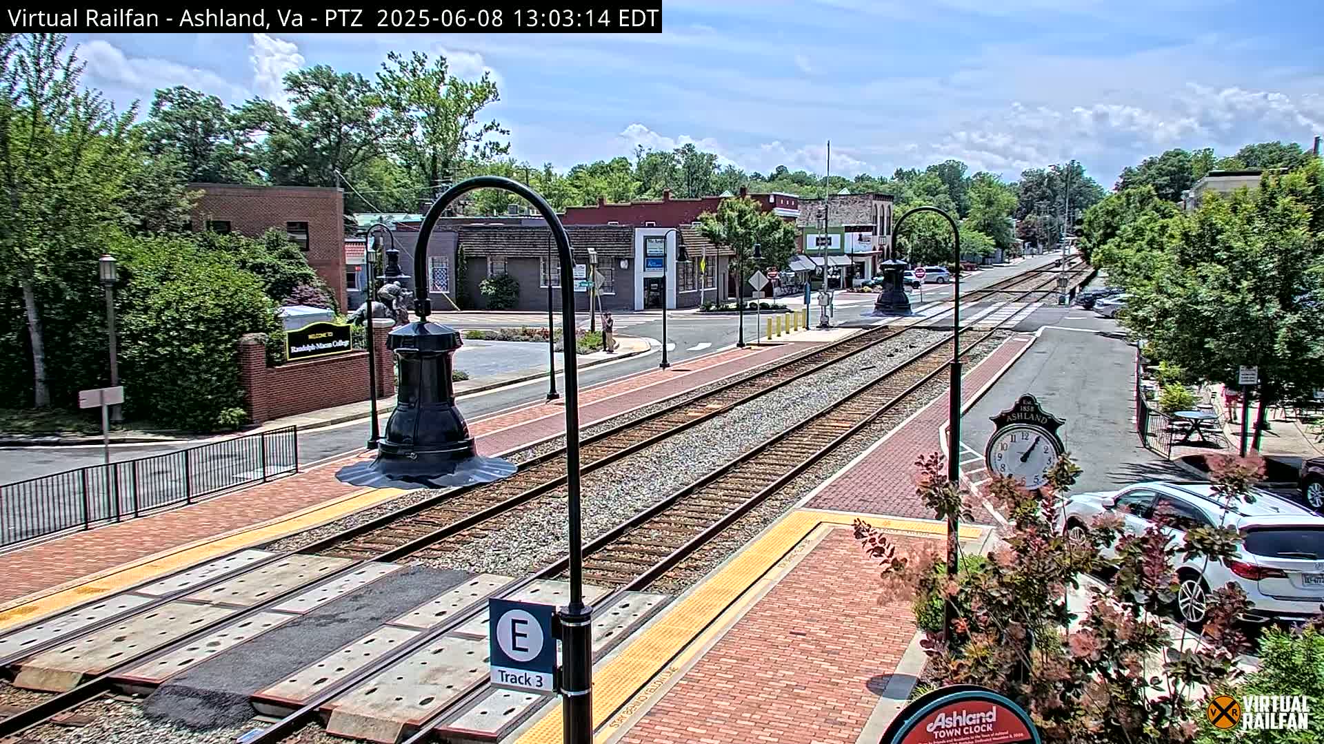 A sunny day reveals a train track running through a town center, lined with brick sidewalks, buildings, and lush greenery.