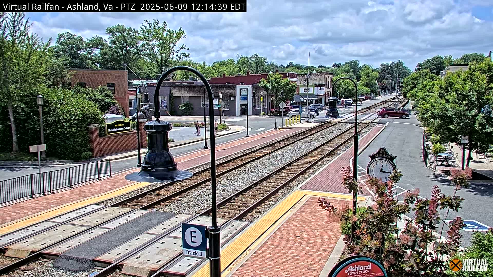 A train track runs through a town on a partly cloudy day, passing by brick buildings and landscaping.
