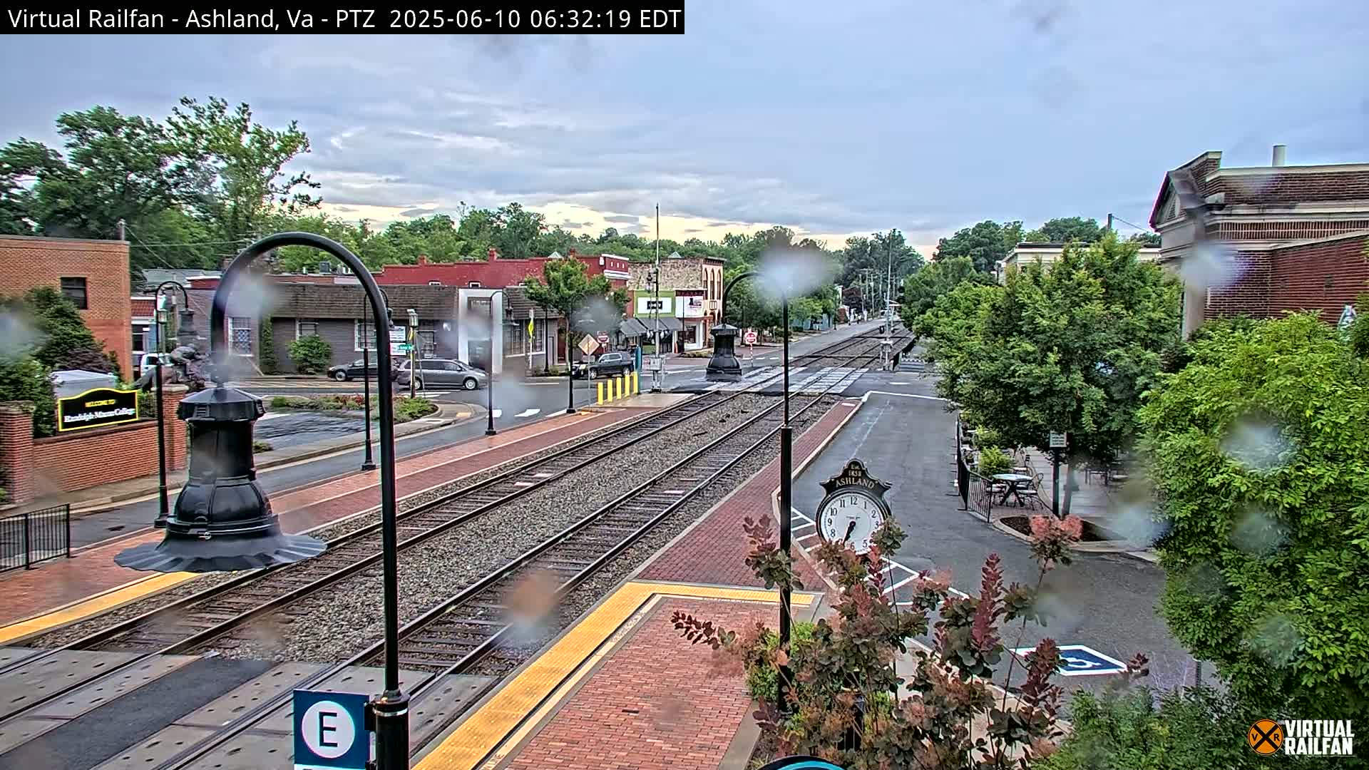 A rain-slicked view of a town's train station and surrounding streets, with brick sidewalks and buildings, and lush green trees.