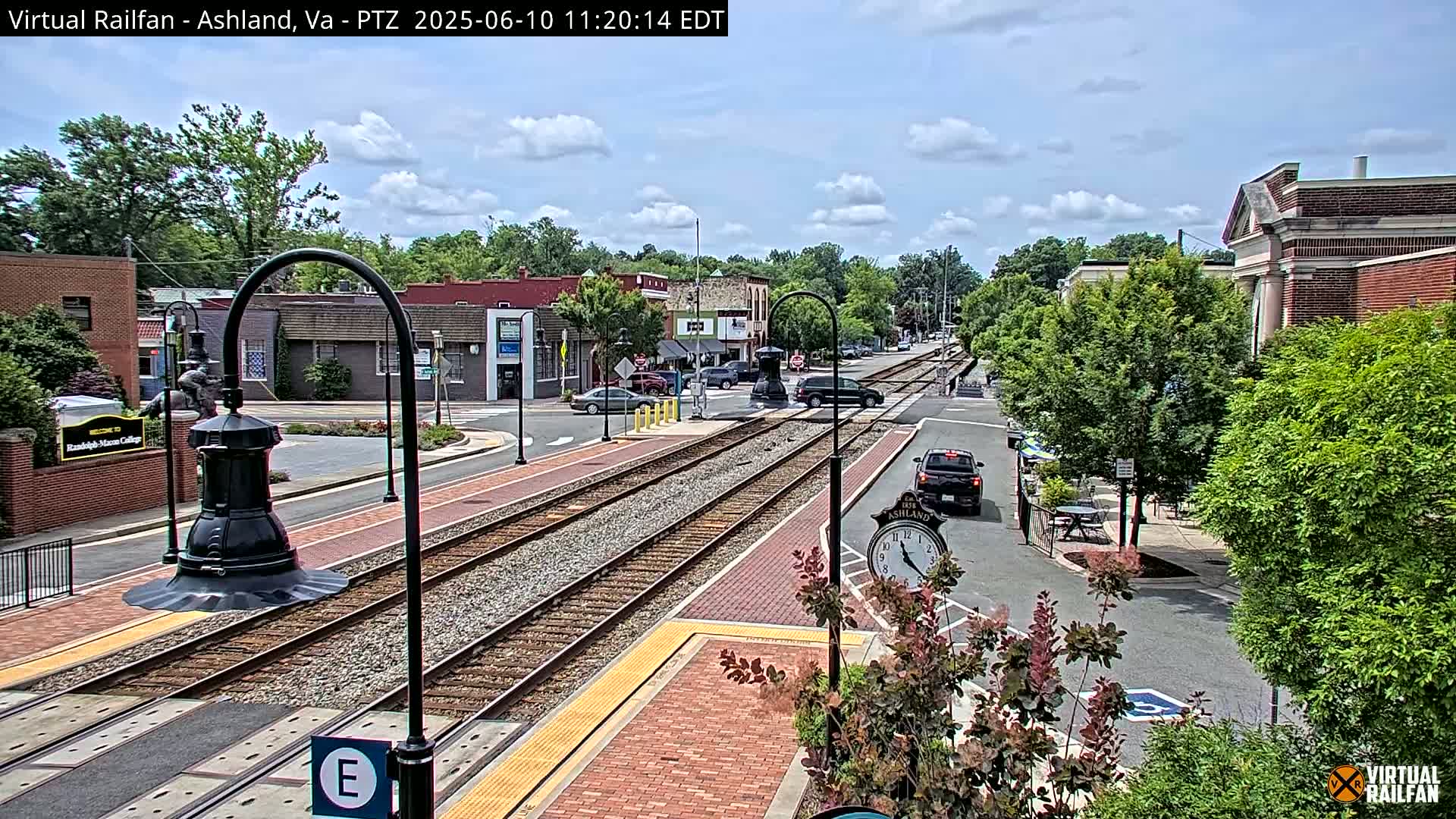 A partly cloudy day reveals a small town's train tracks running through its streets, bordered by brick sidewalks and buildings, with several cars visible and lush green trees lining the sides.