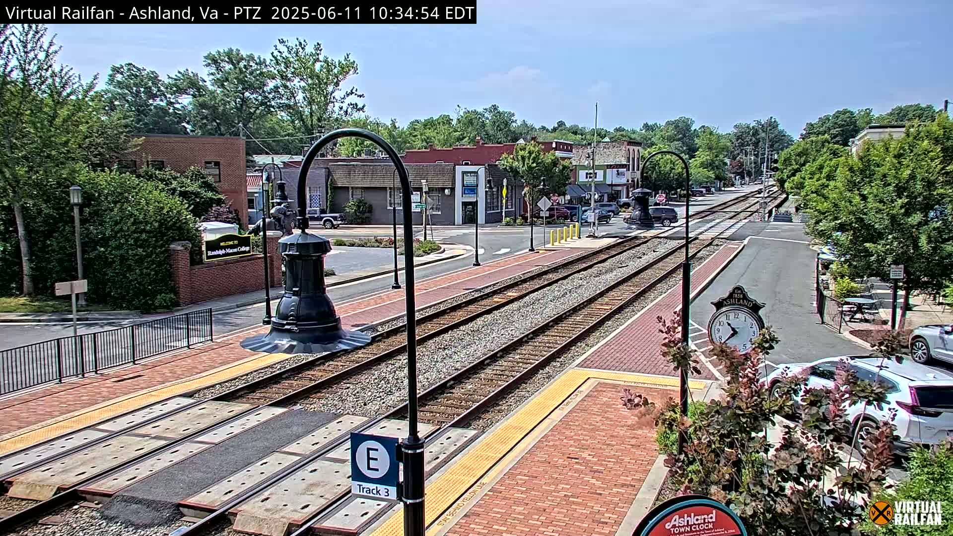 A sunny day in a town features train tracks running through the center of a brick-paved street, with buildings and lush green trees lining the sides.