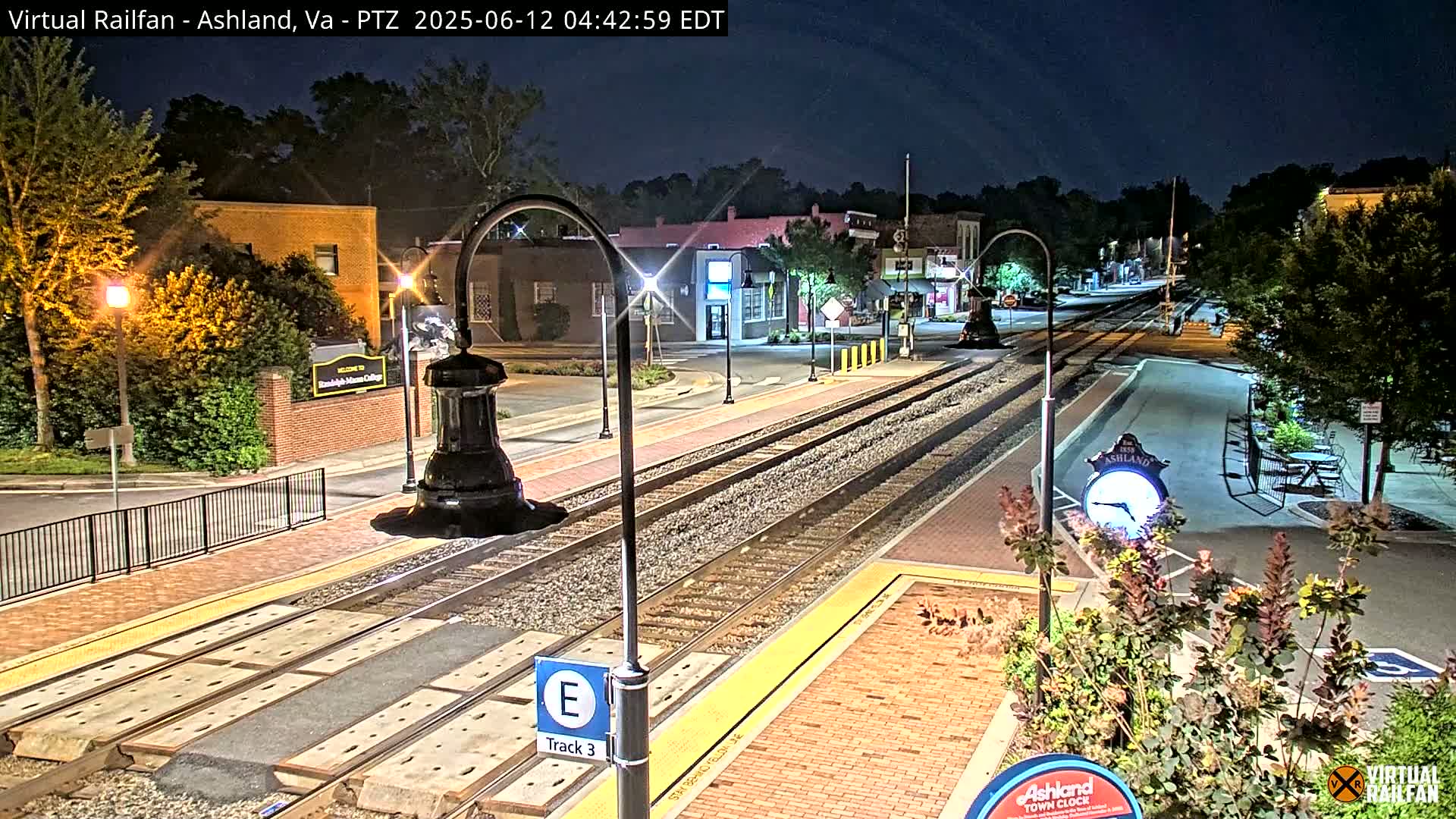 A nighttime view of a train station platform and tracks, with brick pathways and streetlights illuminating a small town's streets and buildings under a clear, dark sky.