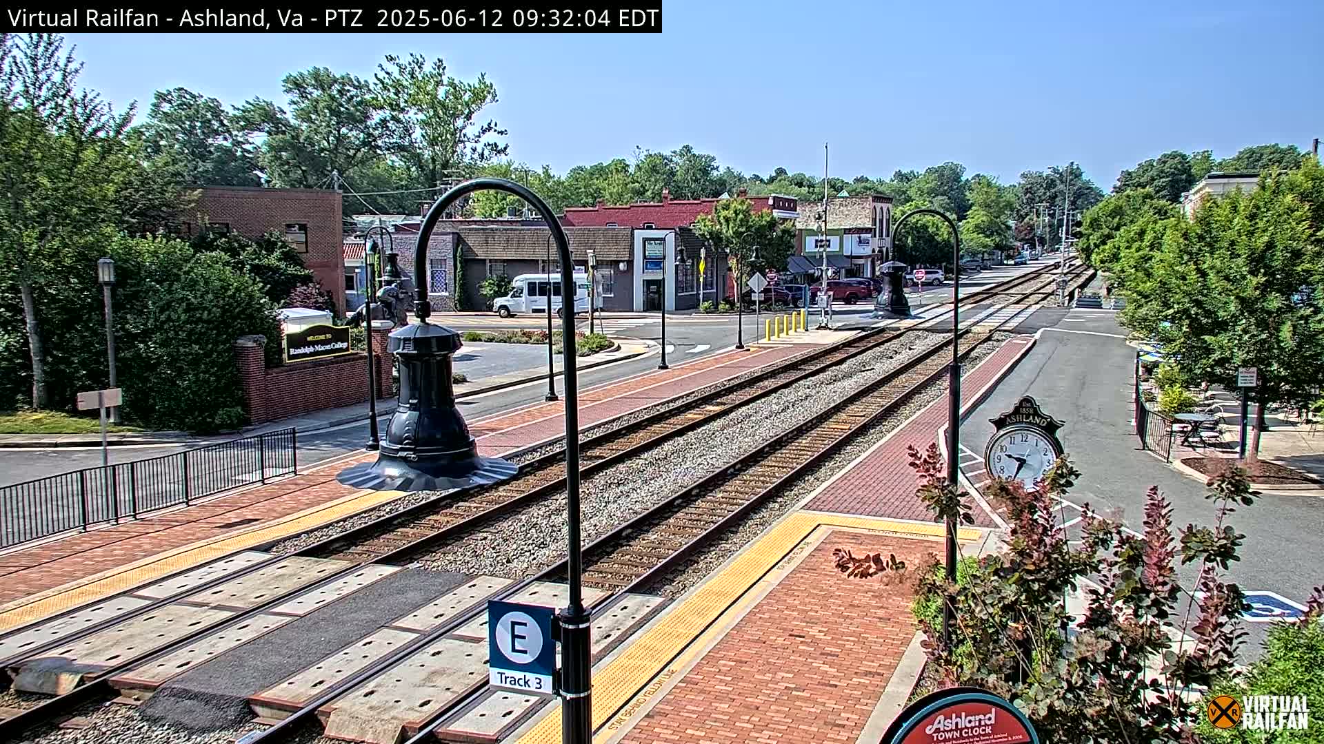 A sunny view of a town's train tracks and surrounding streets, with brick sidewalks and landscaping.