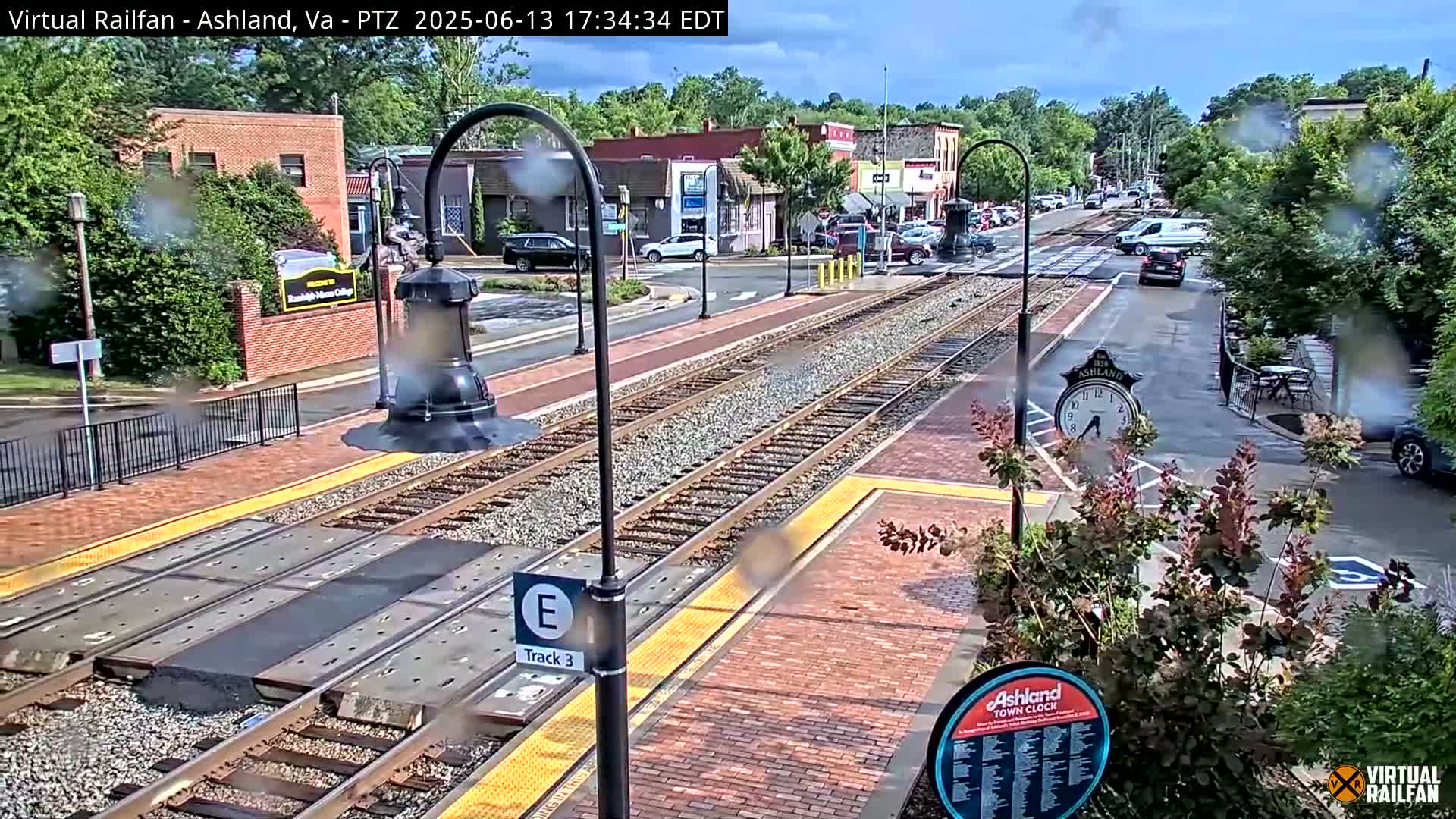 A rain-slicked train station platform with train tracks and a town clock is shown, with several cars parked along a street lined with buildings and trees in the background.