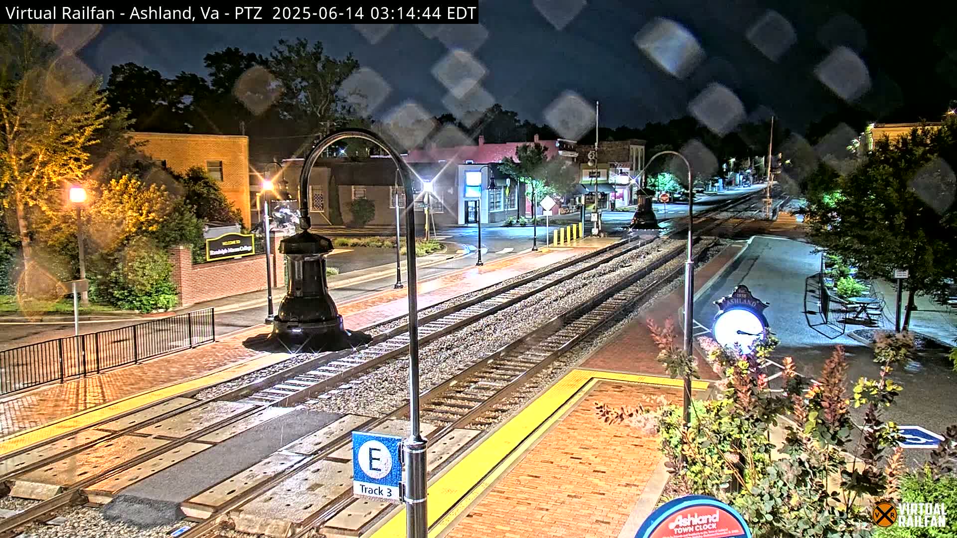 A nighttime view of a train track at a station in a small town, under light rain.