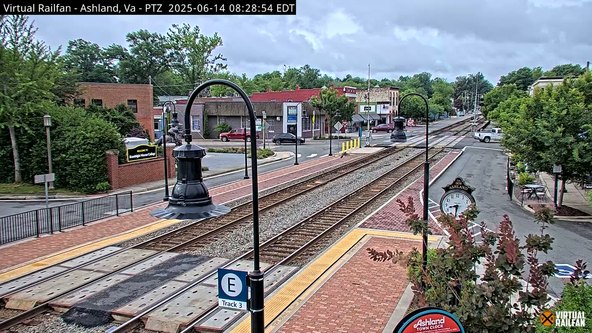 A train track runs through a town's center on an overcast day, passing near brick buildings and landscaping.