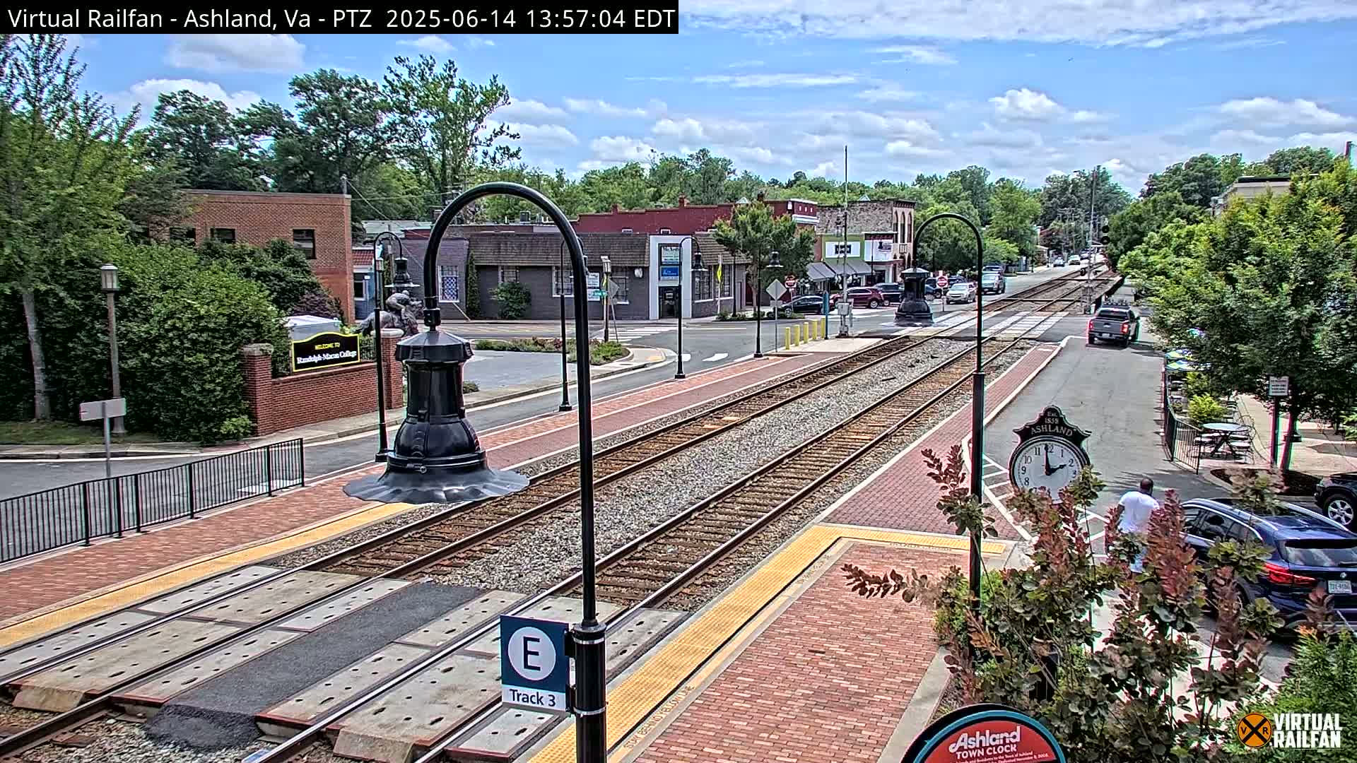 A sunny day view of a town's train tracks and street, with brick sidewalks, buildings, and cars visible.