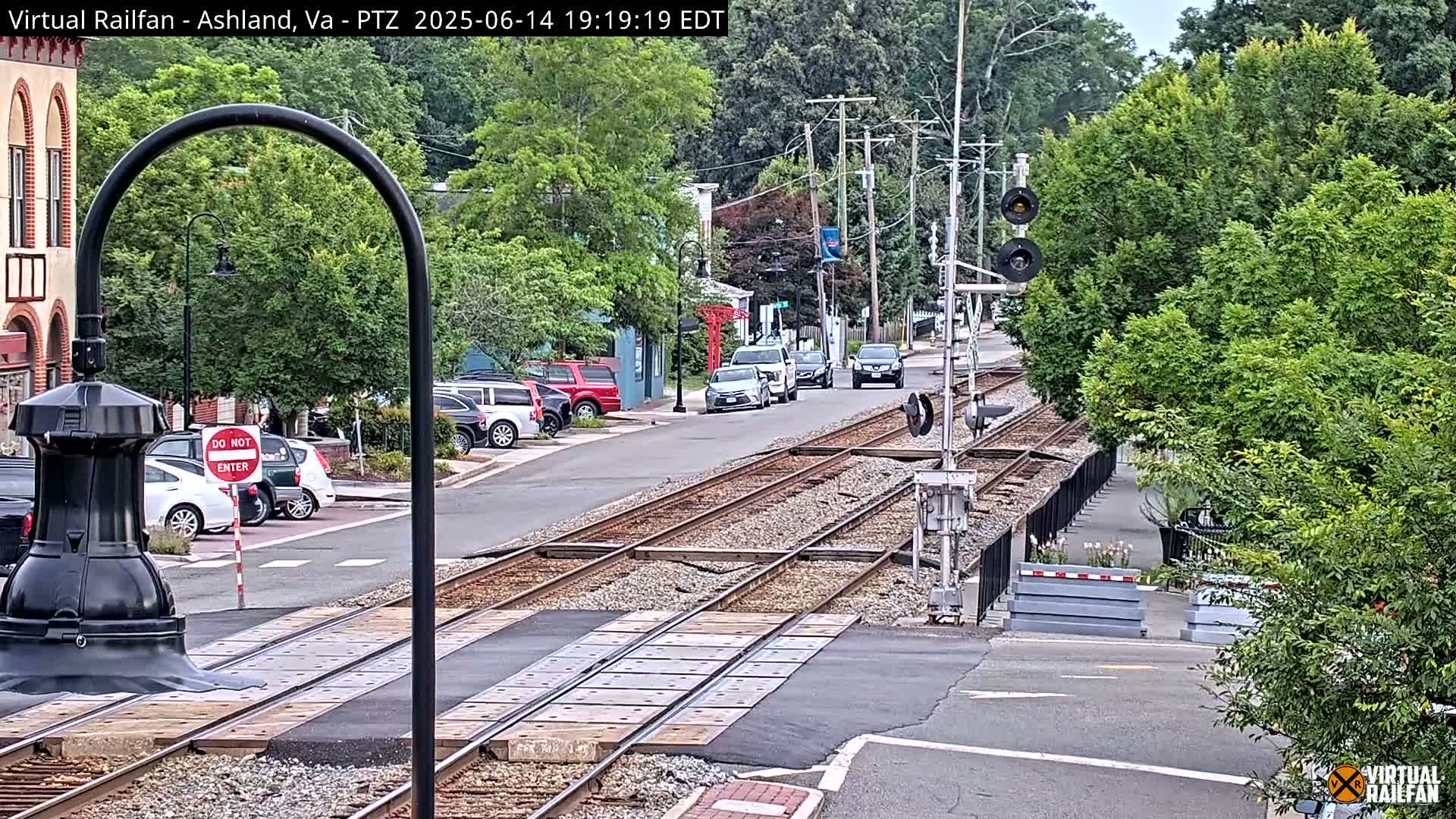 A sunny day view of a town's train tracks and street, with brick sidewalks, buildings, and cars visible.