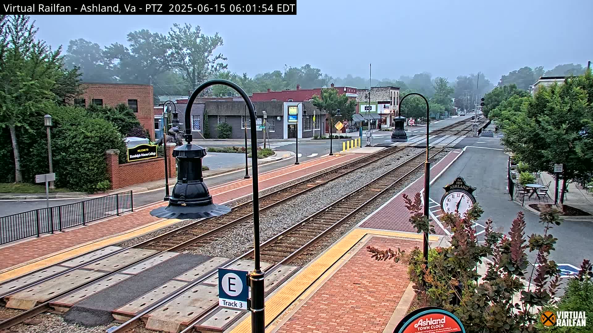 A train track runs through a town square on a foggy morning, with brick walkways and low-lying landscaping surrounding the tracks.