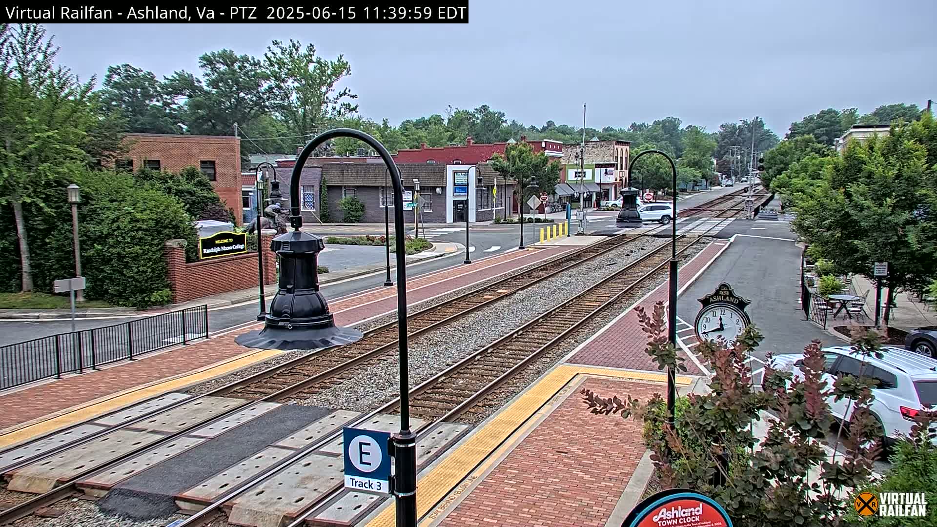 A partly cloudy day reveals train tracks at a station in a small town, with brick sidewalks and buildings lining the street.