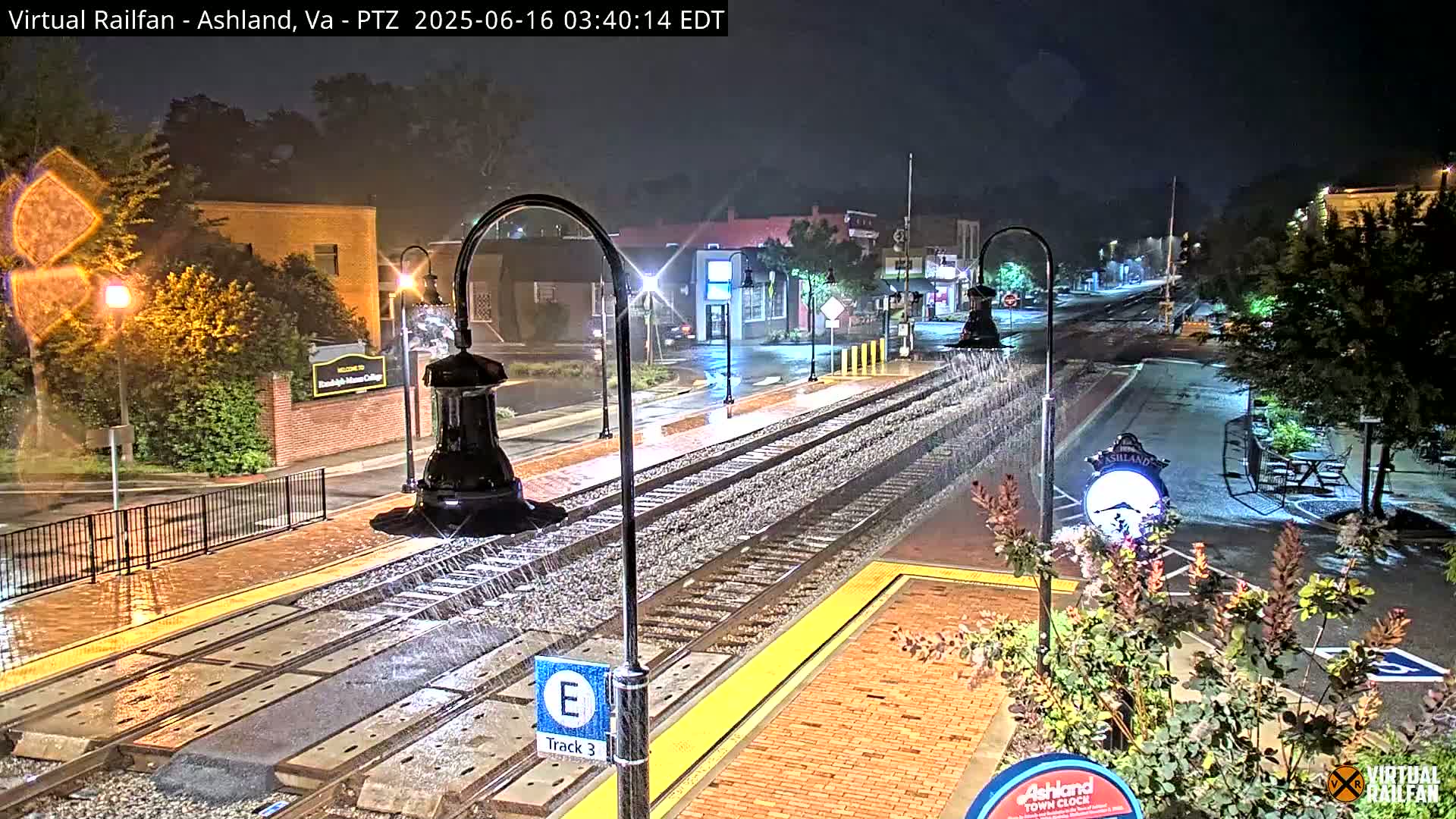 Rain falls on a nighttime scene of train tracks and a town square with streetlights and buildings.