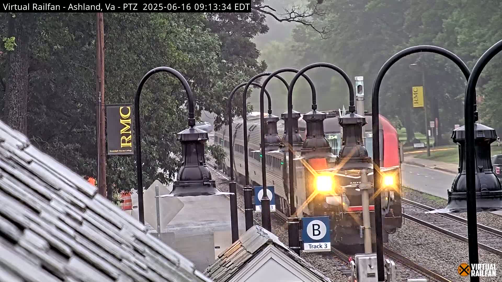 A passenger train approaches a station on a rainy day, passing under several ornate lampposts.