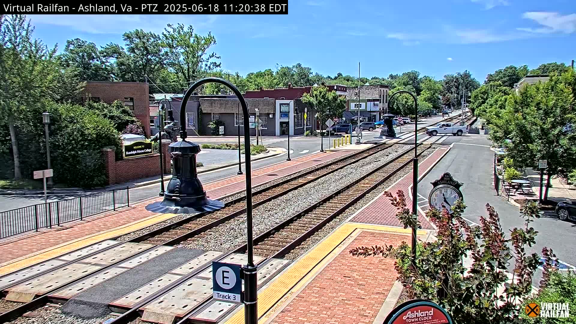 A sunny day reveals a train track running through a town square, with buildings and green trees lining the street.