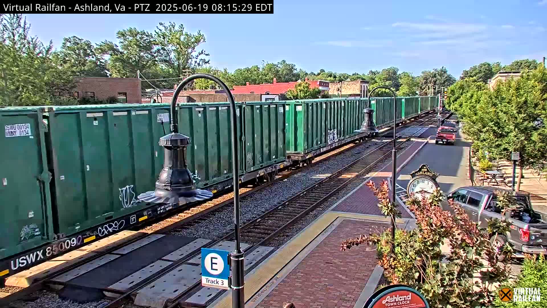 A long freight train with green containers passes by a town's train station on a sunny day.