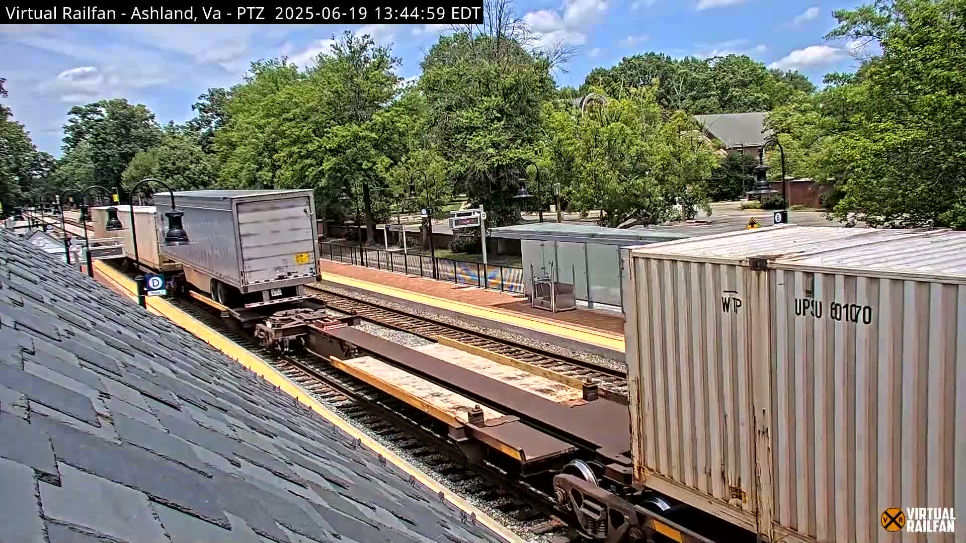 A train carrying two shipping containers and a flatbed trailer passes through a train station on a partly sunny day.