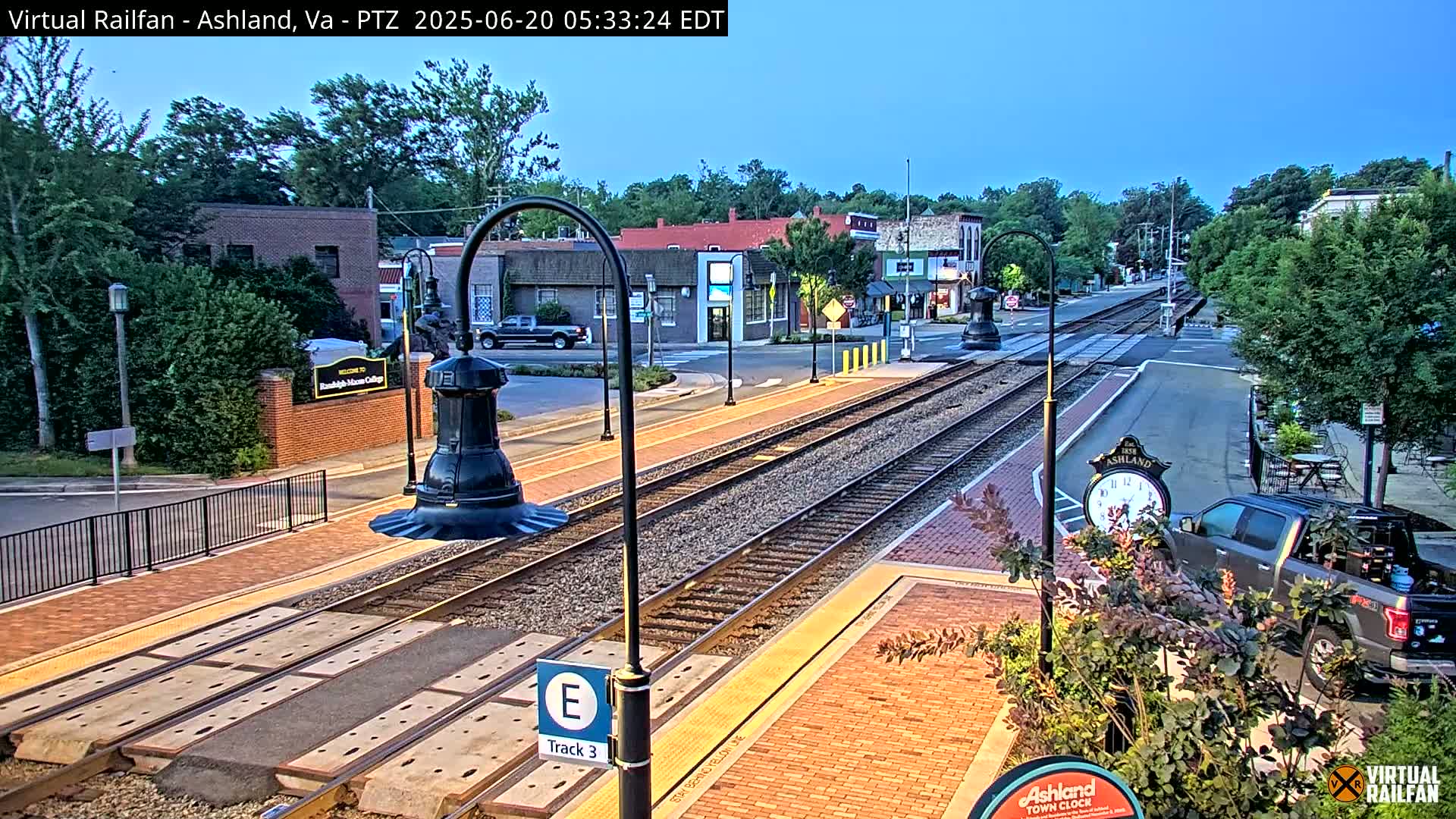 A train track runs through a town square at dawn, with several buildings and a clock visible.