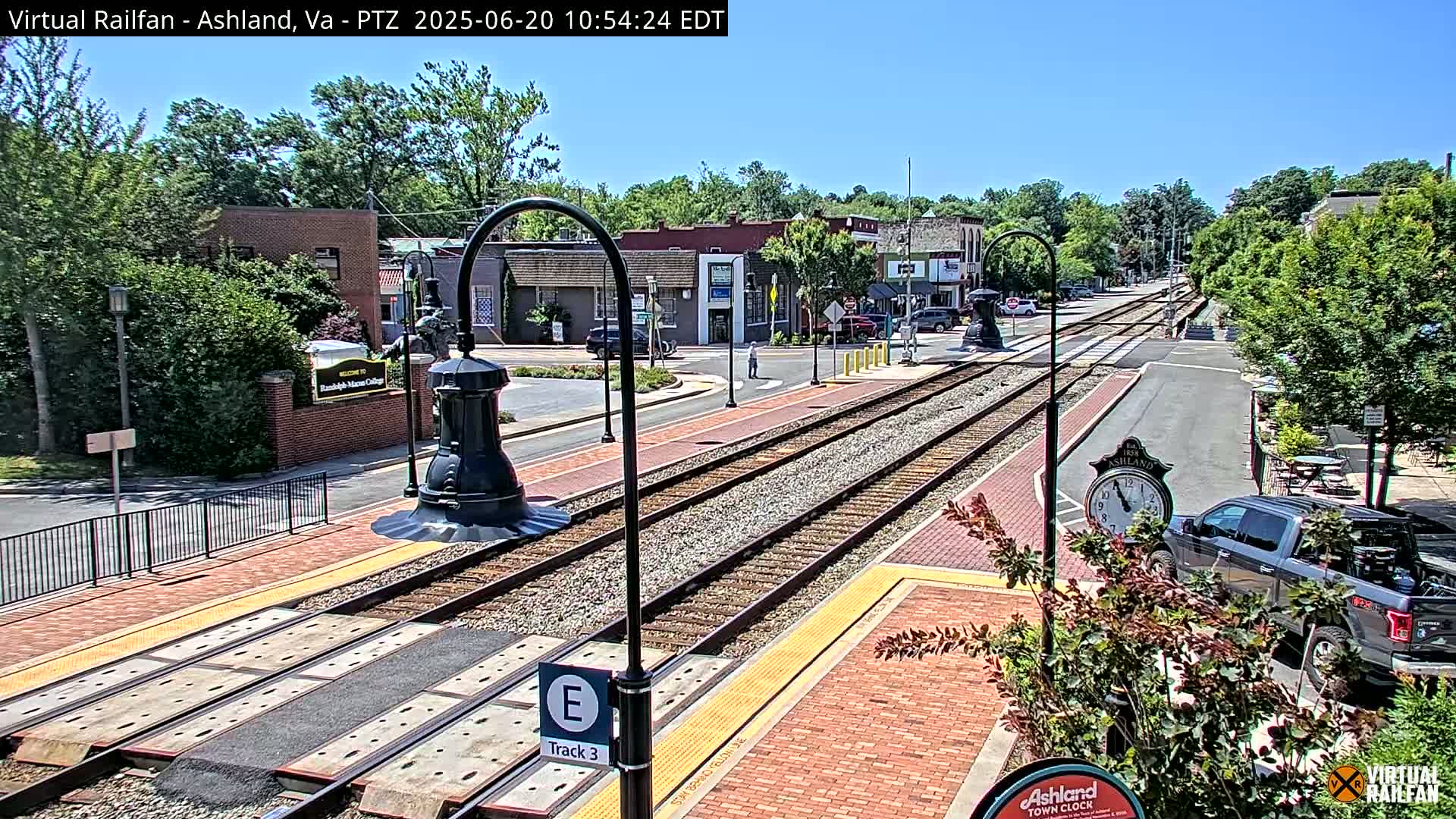 A sunny day shows a view of train tracks running through a town, with brick sidewalks, buildings, and landscaping visible along the sides.