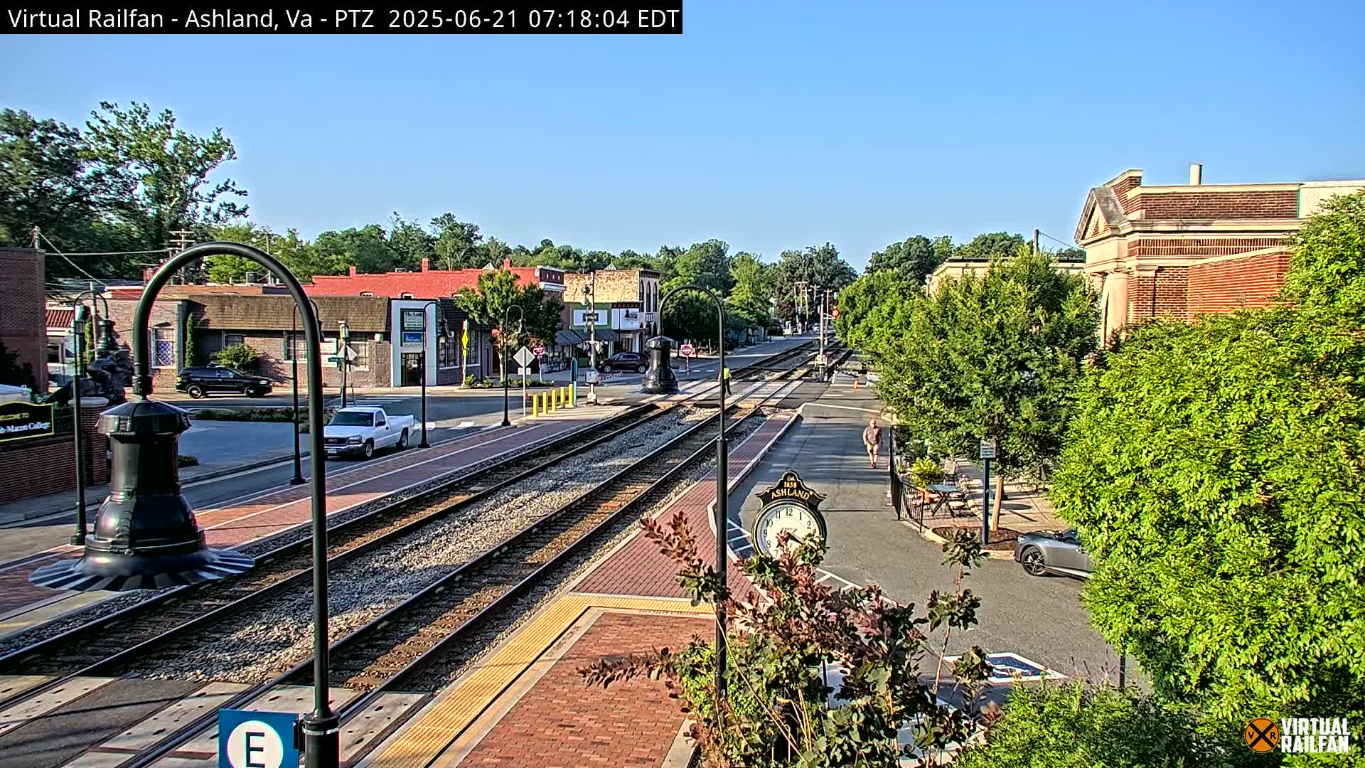 A sunny day in a small town, featuring train tracks running through the center of a street lined with buildings and lush green trees.