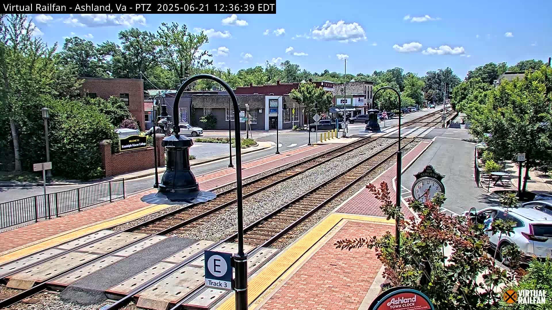 A sunny day shows train tracks running through a town's center, with buildings and trees lining the streets.