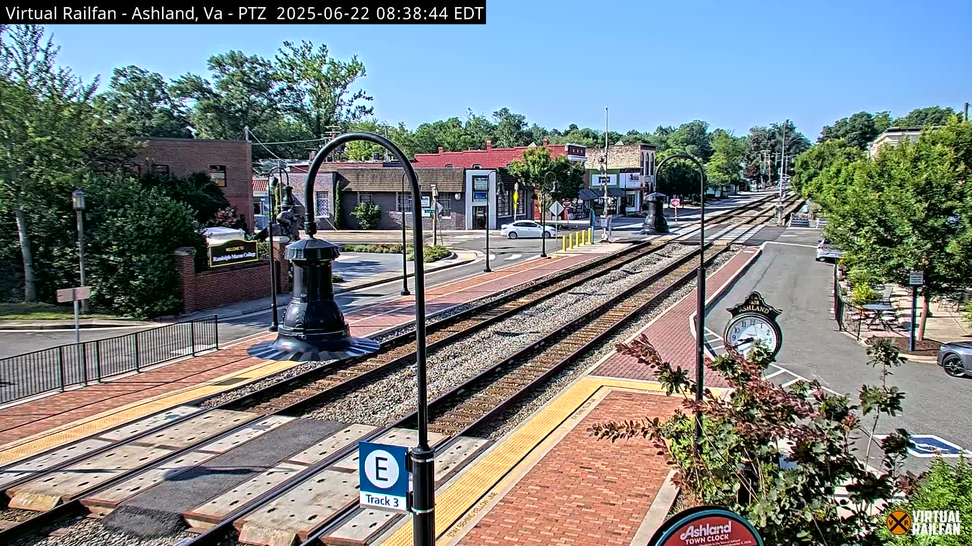 A sunny day reveals a train track running through a town center, bordered by brick sidewalks, buildings, and lush green trees.