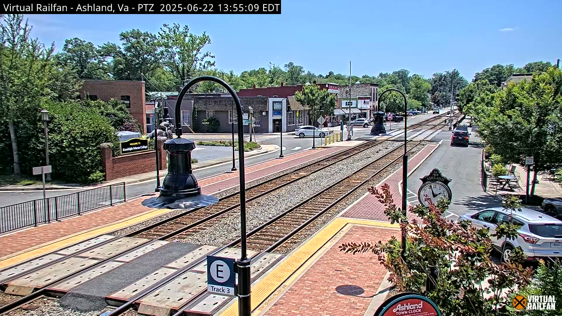 A sunny day view of a town's train tracks, with several cars parked along the side and buildings lining the street.