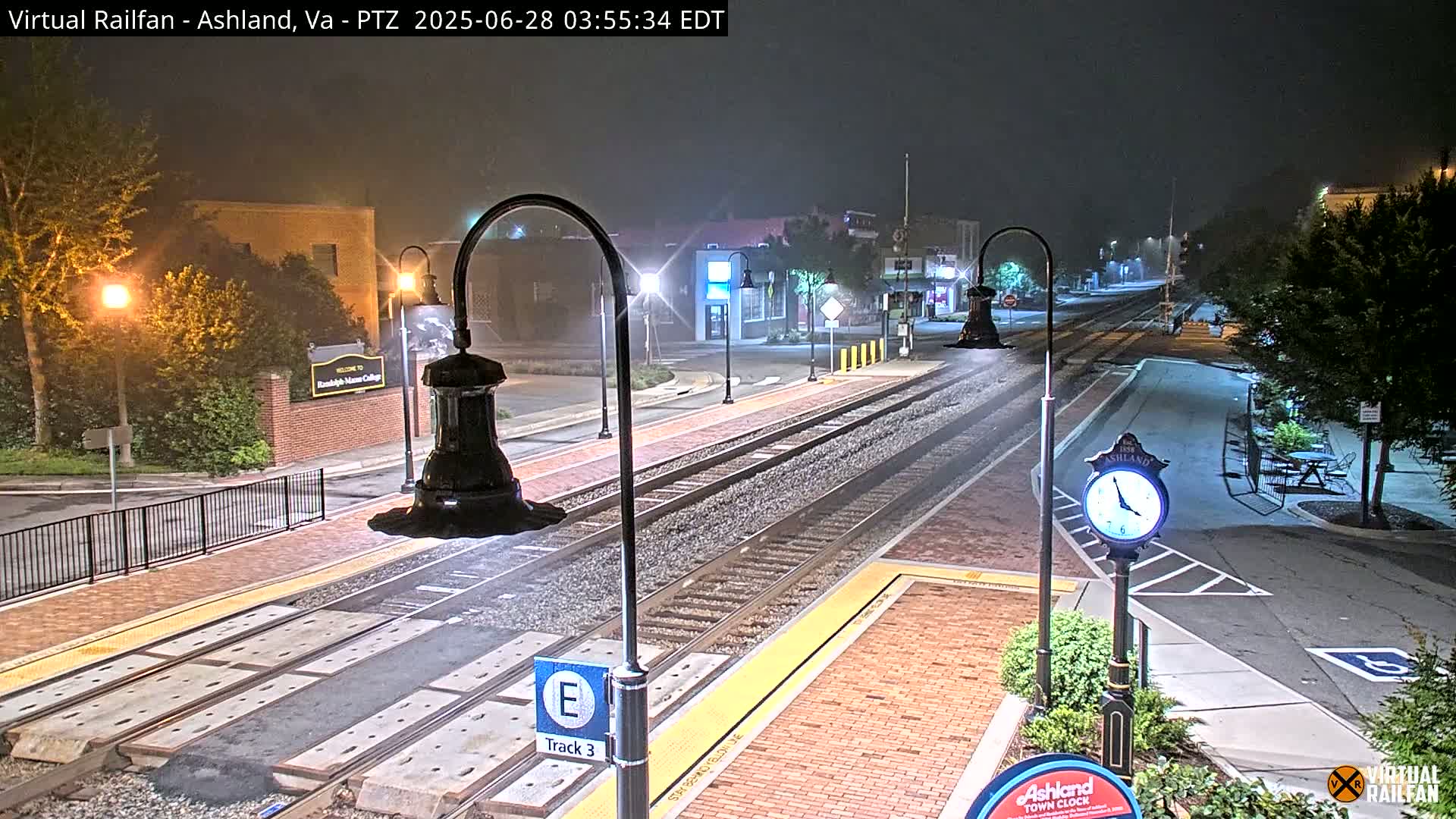 A nighttime view of a train station platform with tracks and streetlights, under hazy conditions.