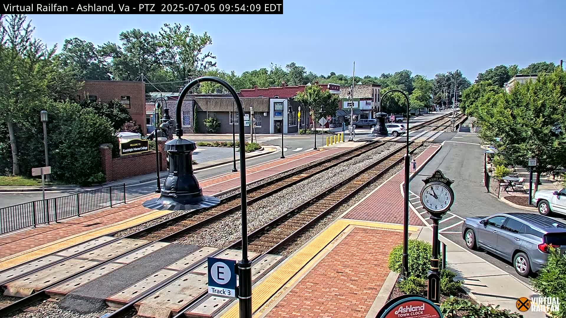 A sunny day reveals a train track running through a town's brick-paved streets, lined with buildings and lush green trees.