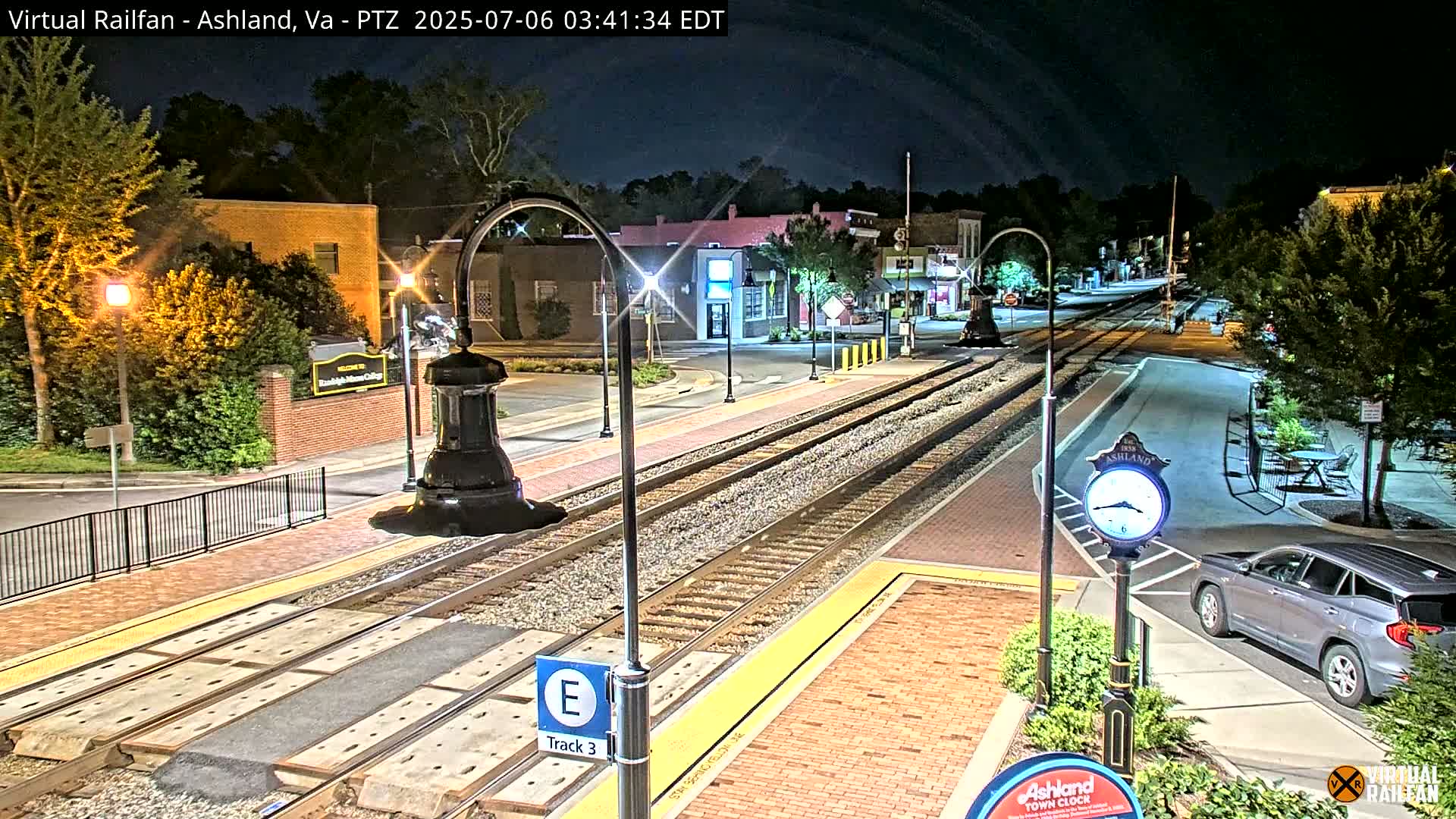 A nighttime view of a train track and platform in a town, with streetlights illuminating the area and a car parked nearby.