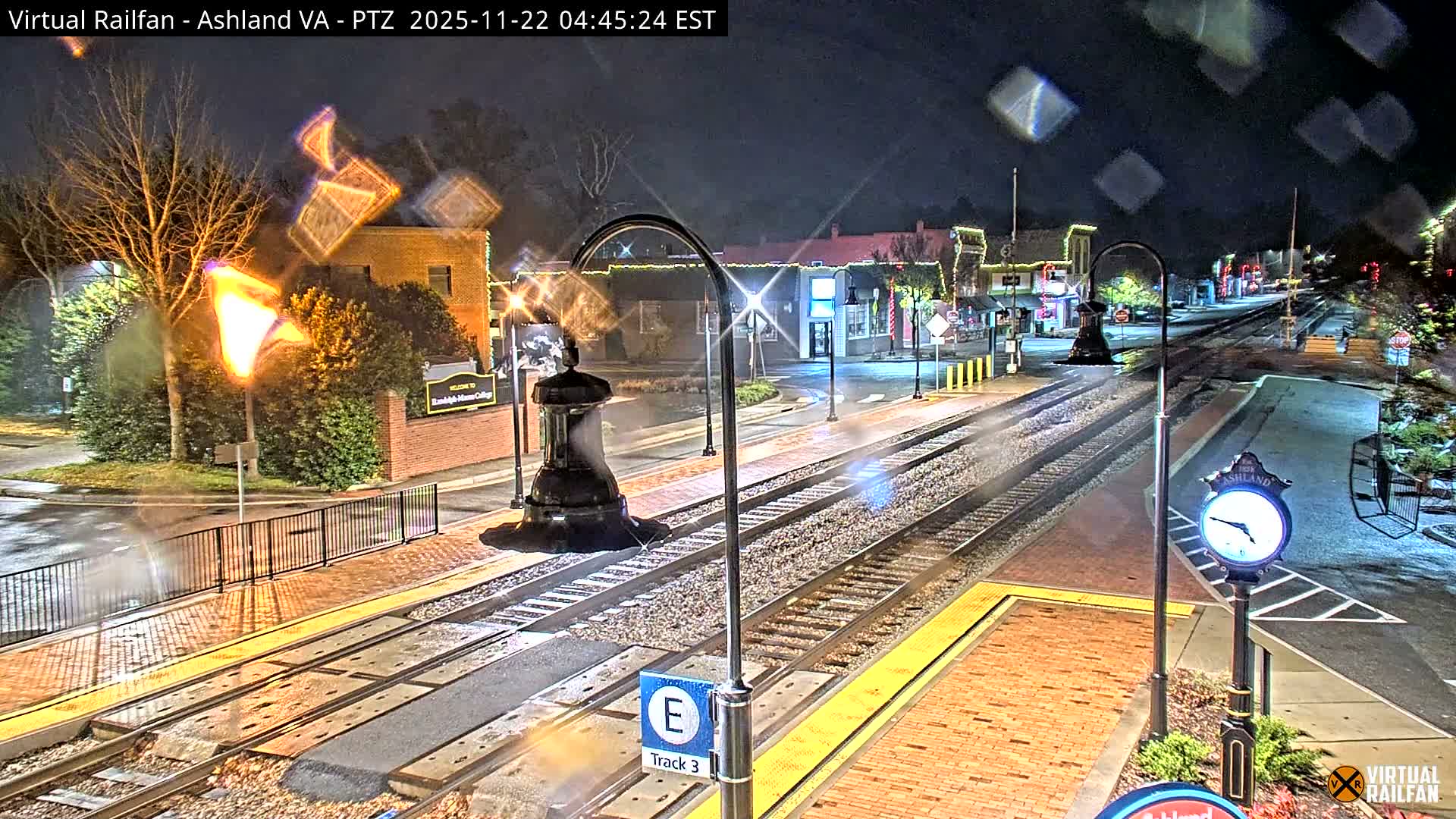 A nighttime scene of a small-town train station features wet tracks and platforms, illuminated by streetlights and buildings adorned with festive lights, indicating recent rain.