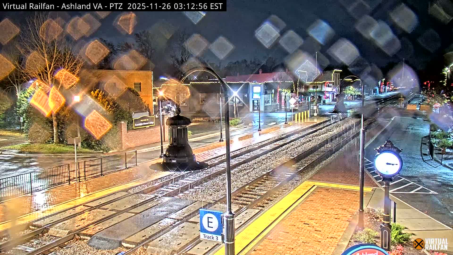 A nighttime scene depicts a wet train station and tracks running alongside a town street lined with illuminated buildings, all under rainy conditions, viewed through a rain-streaked lens.