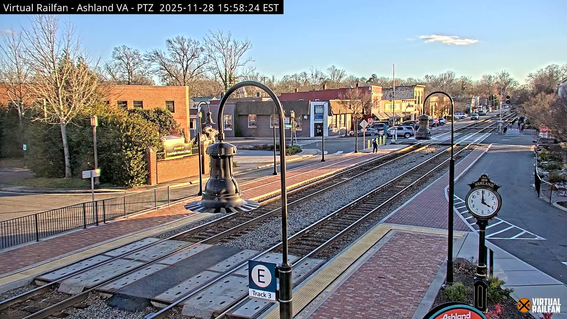 A clear, sunny day illuminates a small town where railroad tracks run through the center, flanked by brick buildings, parked vehicles, and scattered pedestrians.