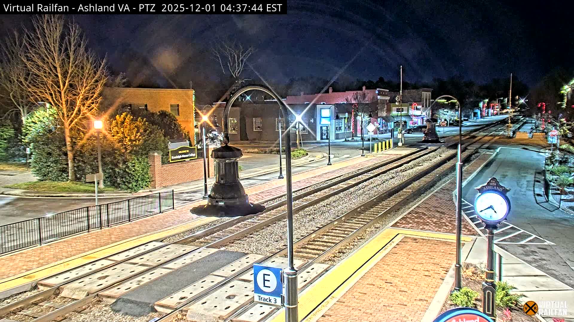 A clear night illuminates a small town scene featuring train tracks and platforms, surrounded by brightly lit buildings, bare winter trees, and festive decorations.