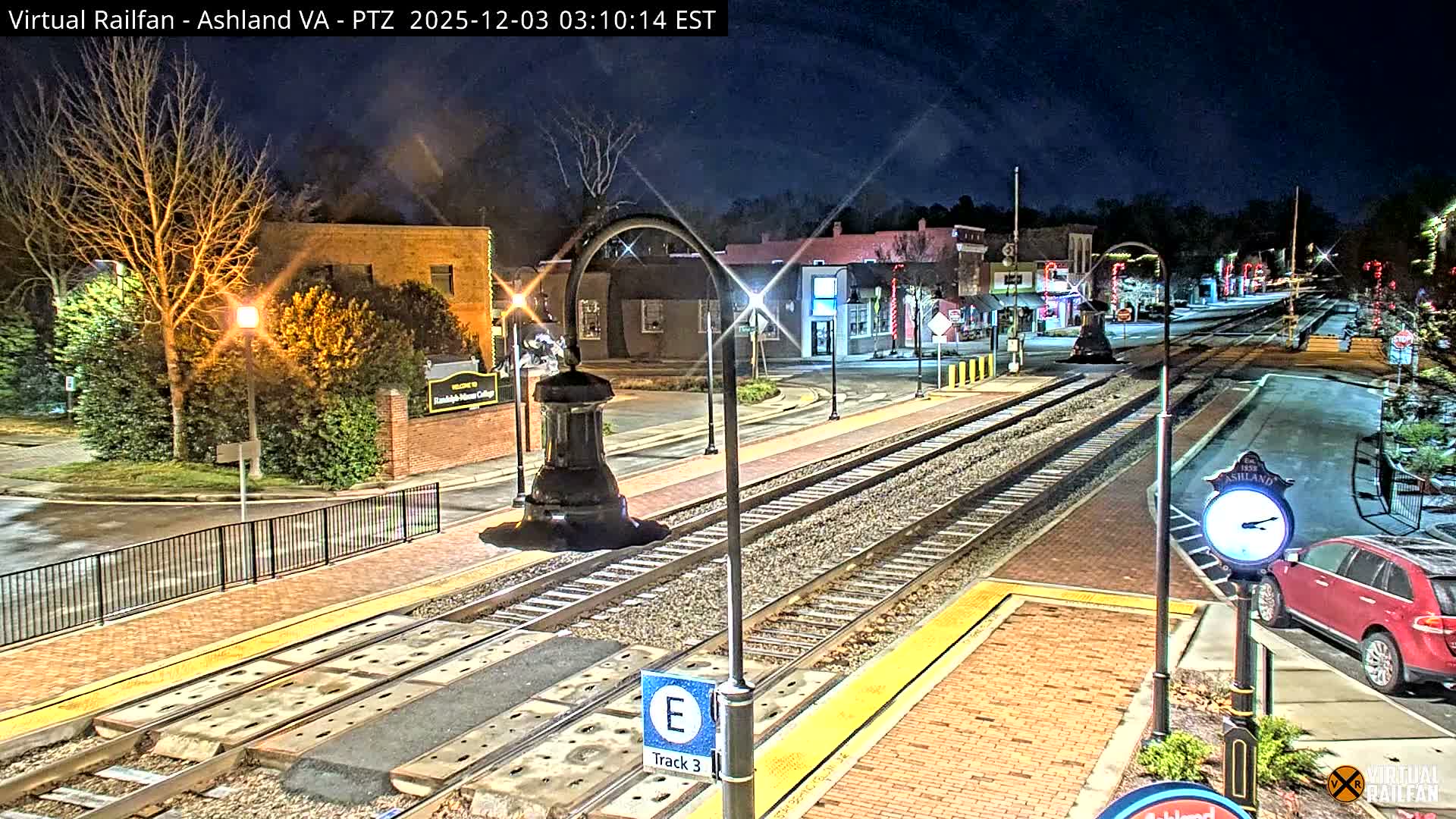A well-lit train station with multiple tracks cuts through a small town featuring buildings and bare trees, all under a clear night sky.