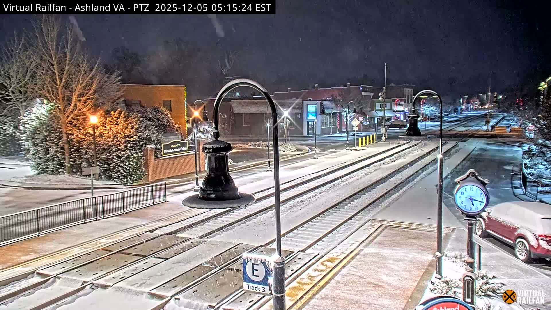 A nighttime view of a snow-covered town shows illuminated train tracks running through a street lined with buildings and sparse trees, under an actively snowing sky.