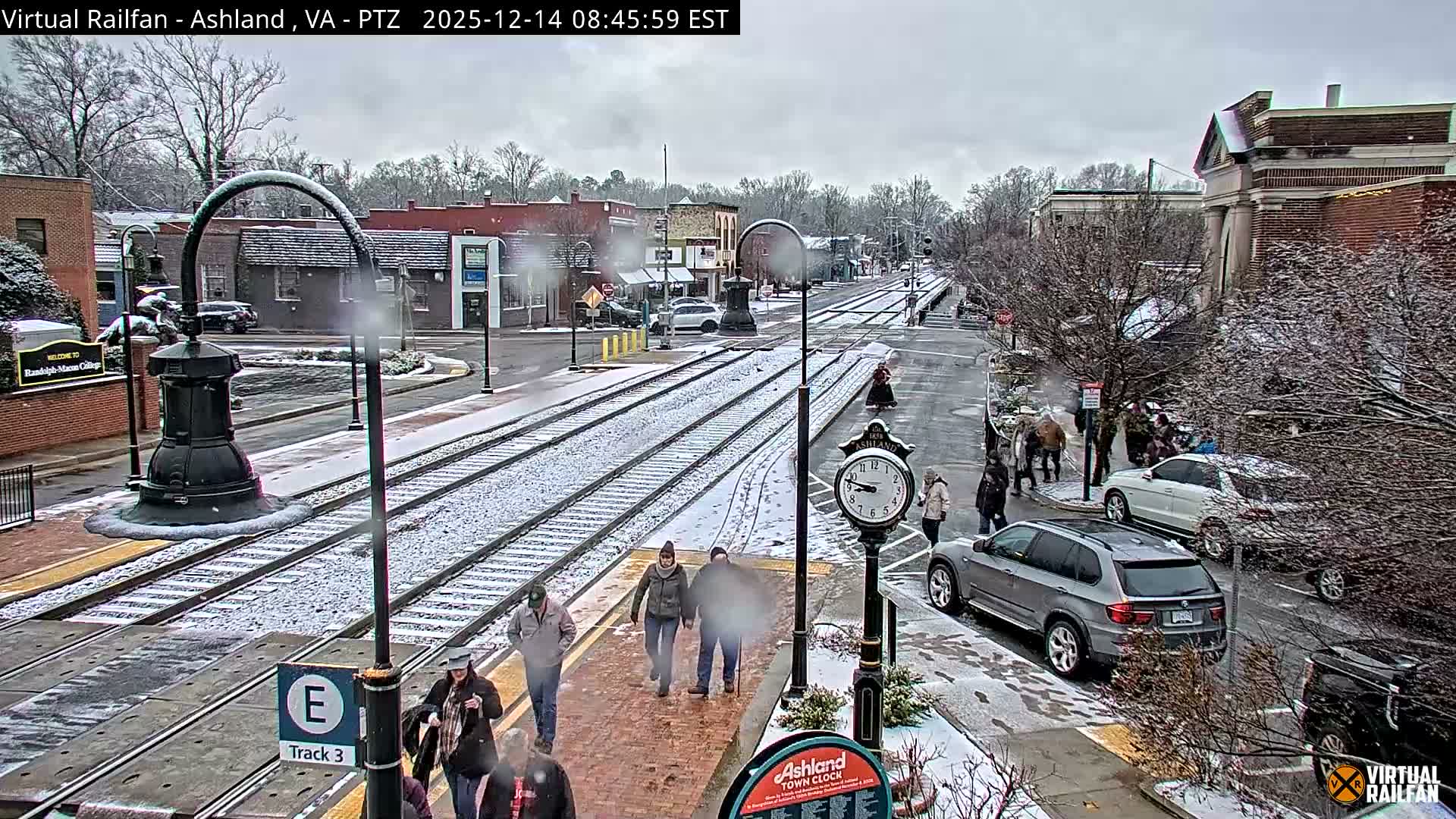 A nighttime view of a snow-covered town shows illuminated train tracks running through a street lined with buildings and sparse trees, under an actively snowing sky.
