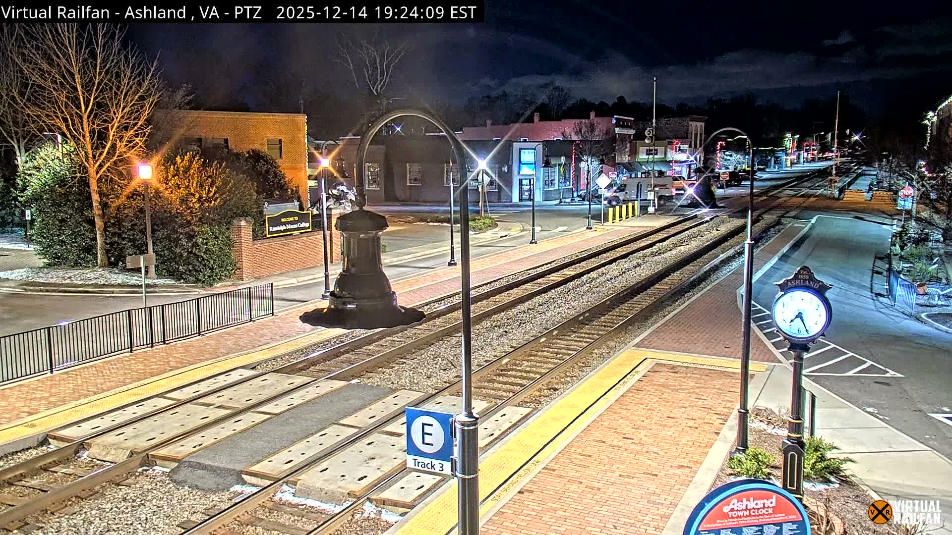 A nighttime view of a snow-covered town shows illuminated train tracks running through a street lined with buildings and sparse trees, under an actively snowing sky.
