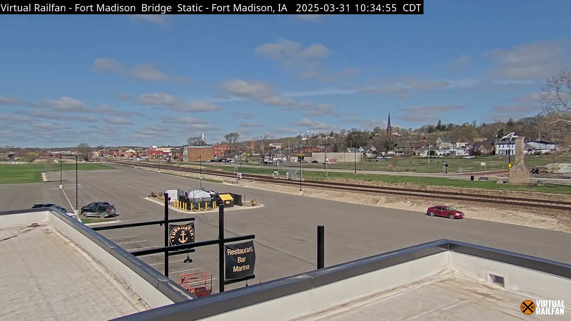 A partly cloudy day reveals a small town viewed from a rooftop parking lot, with railroad tracks separating the lot from the town and a red car visible in the distance.