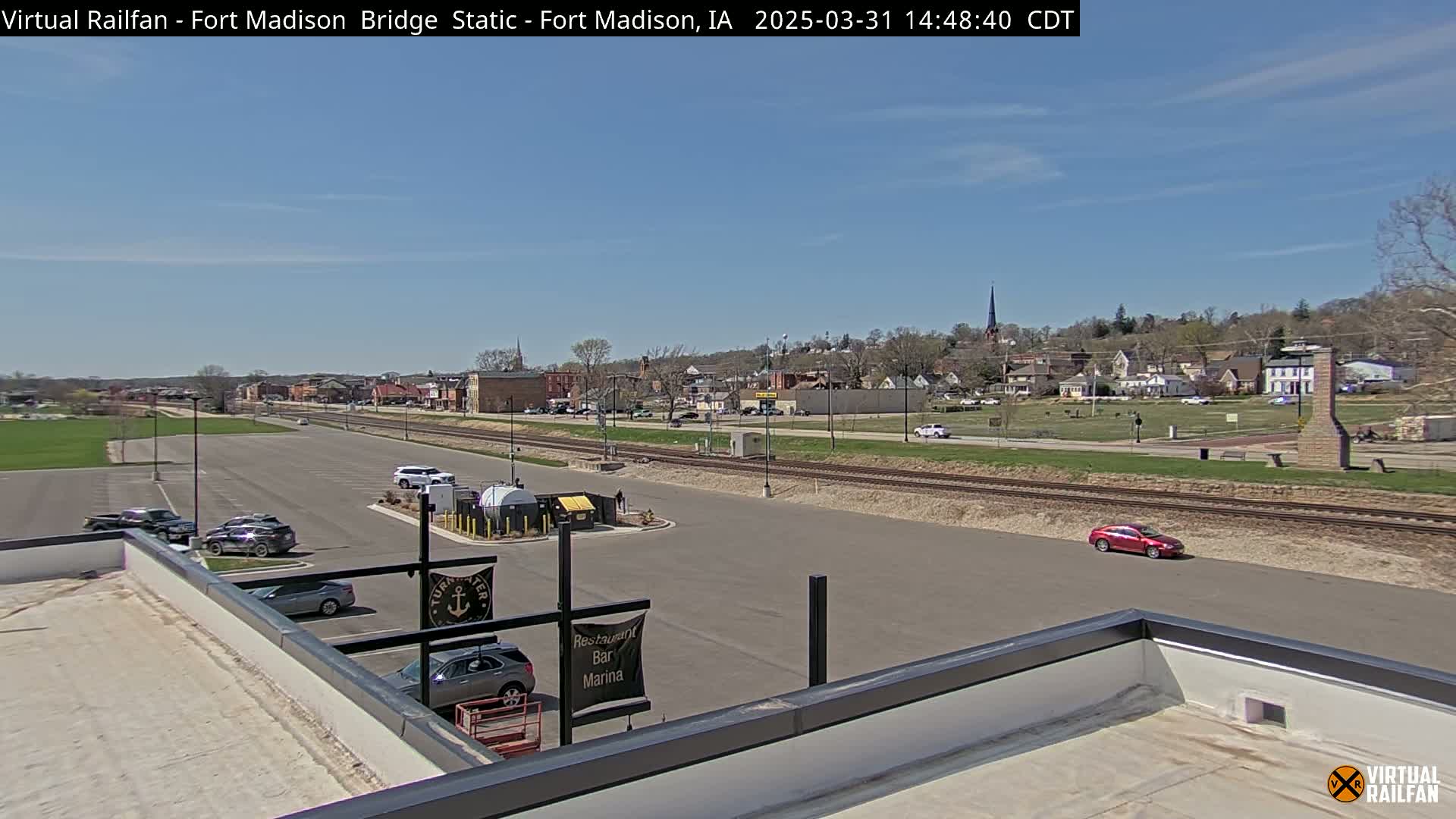 A mostly sunny view of a small town with a river, railroad tracks, parking lot, and a few cars, seen from a rooftop.