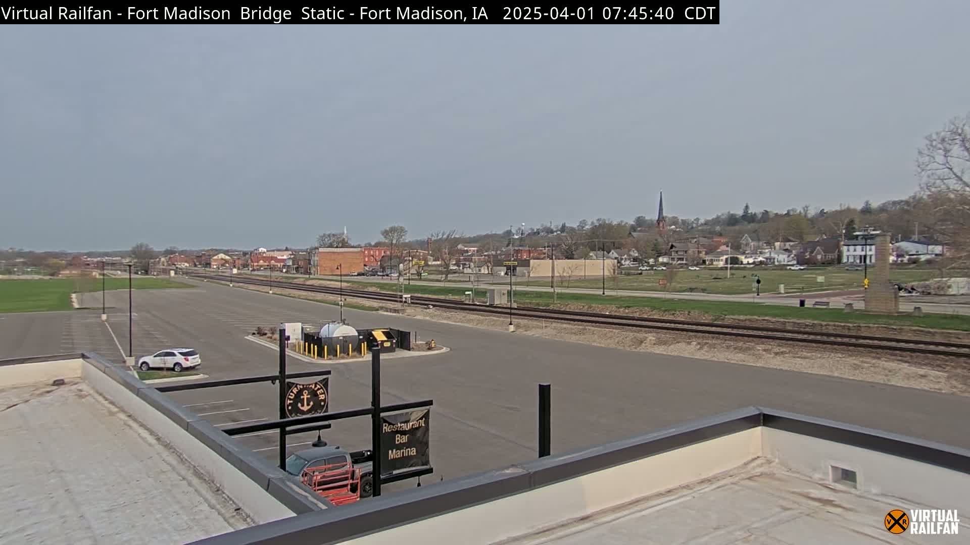 A mostly clear, daytime view of a town, featuring a parking lot, train tracks, and a flat rooftop with a partial view of a building.