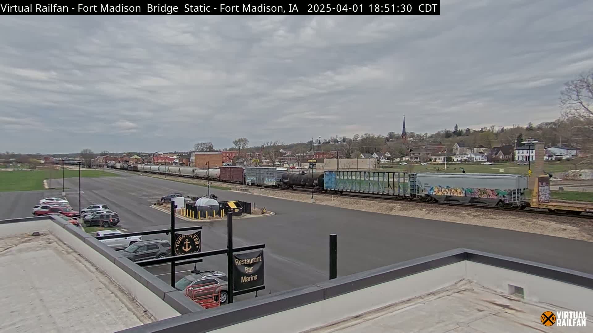 A long freight train passes by a parking lot near a riverfront restaurant under a cloudy sky.
