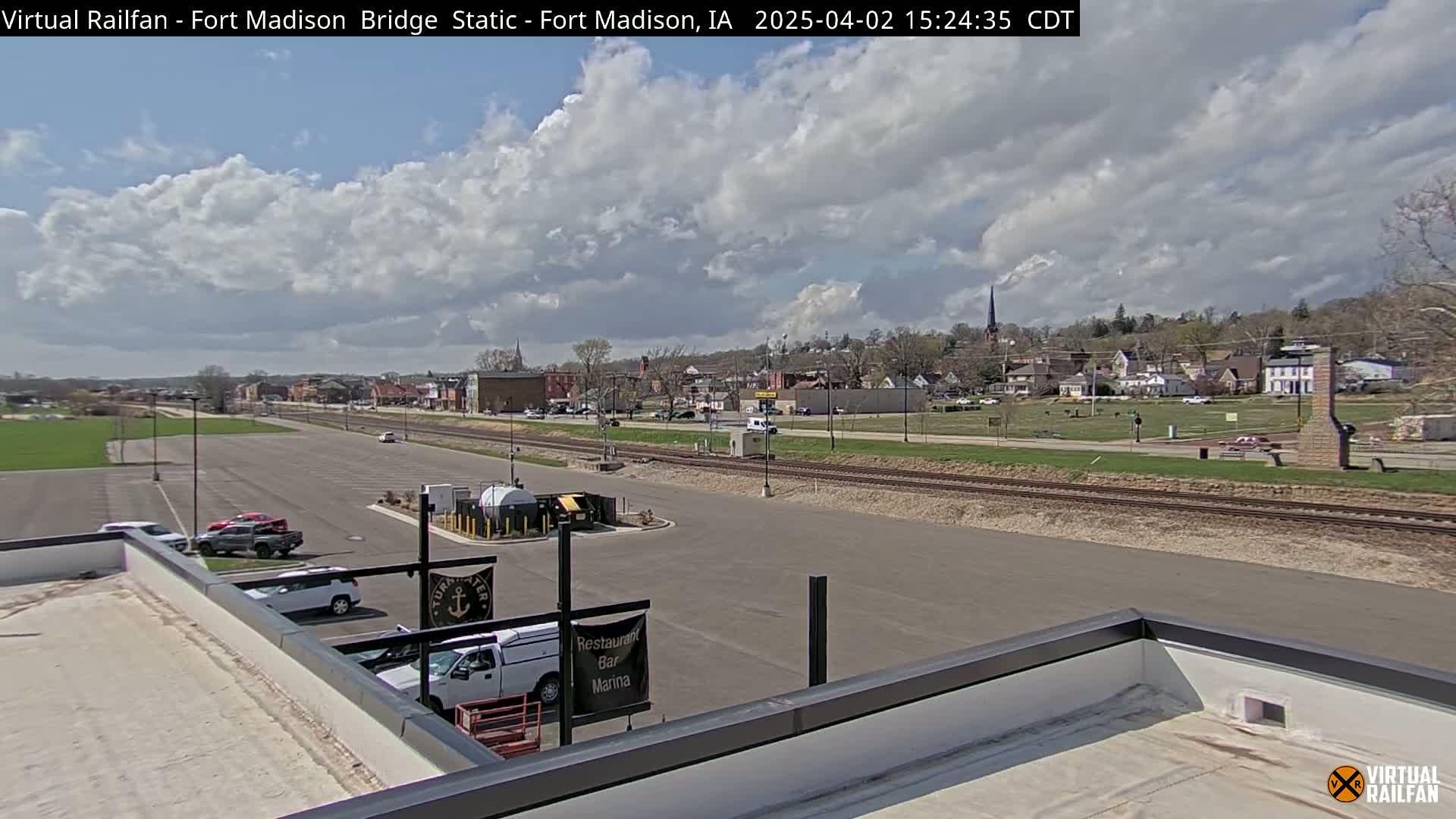 A partly cloudy day reveals a small town viewed from a rooftop, with a parking lot, train tracks, and a few vehicles visible in the foreground.
