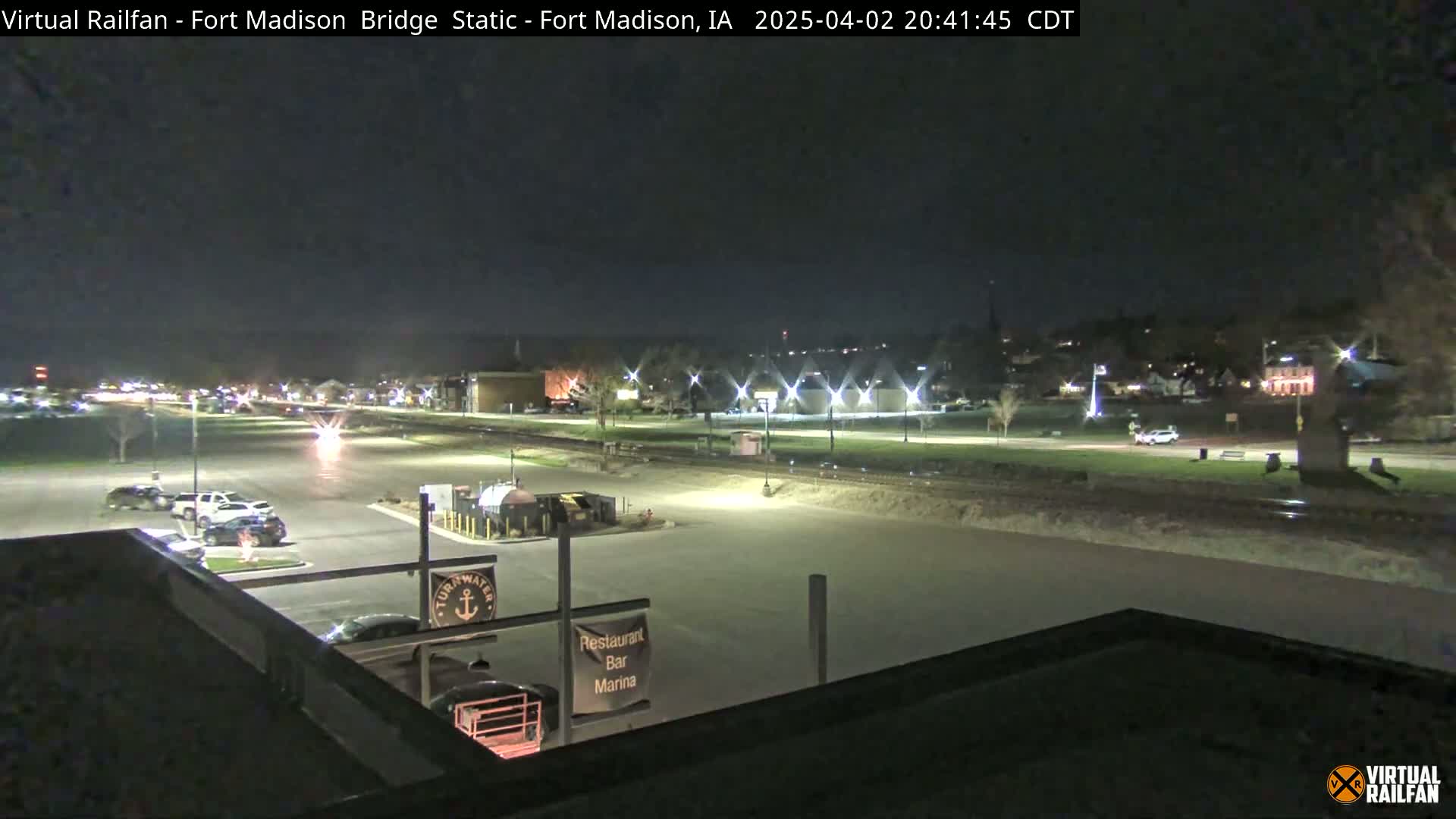 A nighttime, outdoor view of a mostly empty parking lot and a road next to a riverfront, with some cars and buildings visible in the distance under a clear sky.