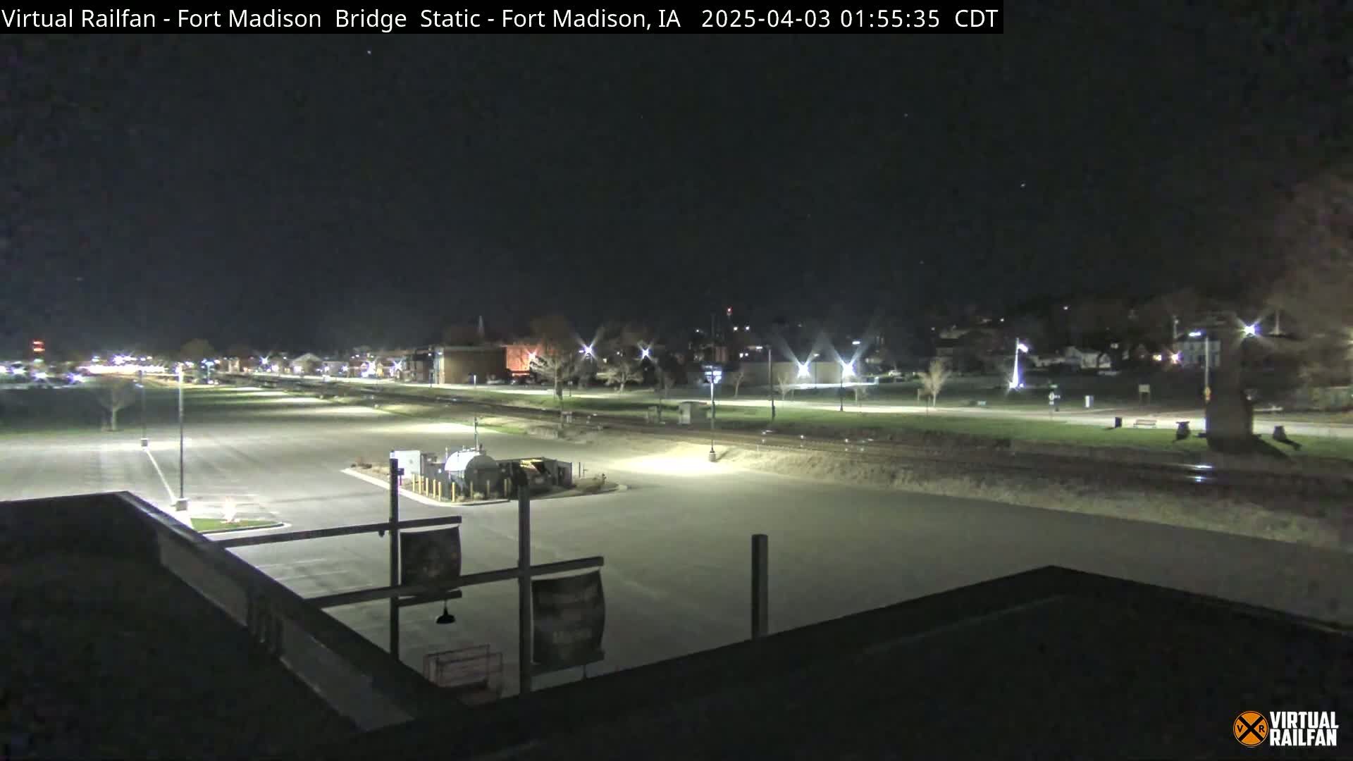 A nighttime view of a mostly empty parking lot and a train track, with some buildings and trees visible in the distance under a clear, dark sky.