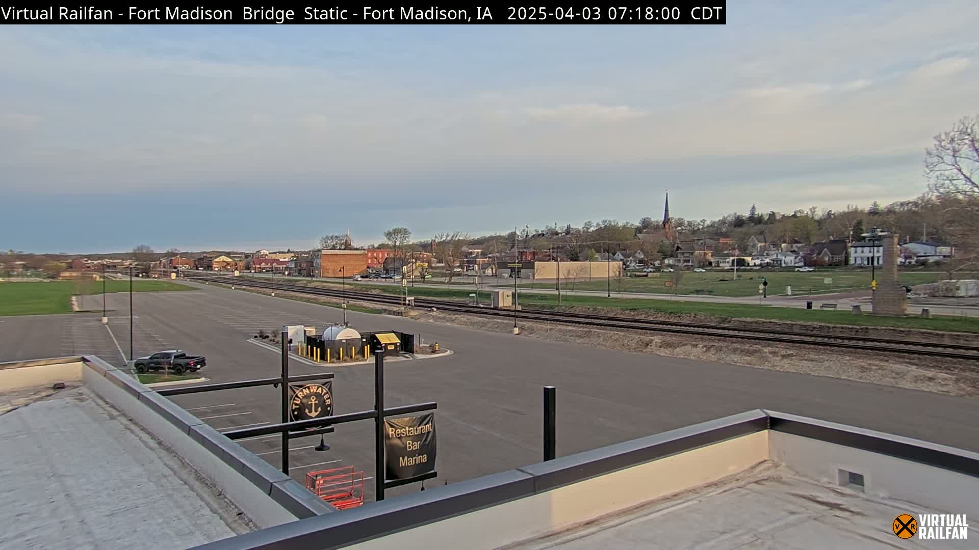 A mostly clear sky over a small town with a parking lot, train tracks, and a body of water in the distance.