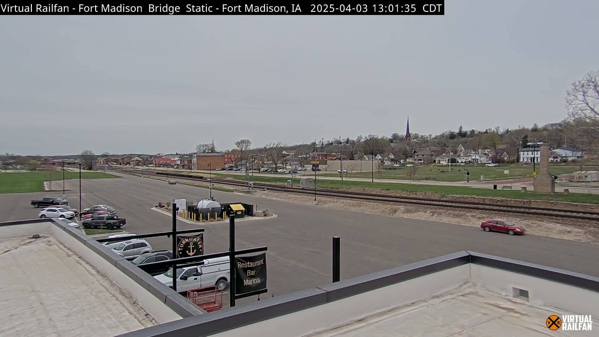 A mostly cloudy day reveals a view of a small town with a parking lot, train tracks, and a few cars, seen from a rooftop.
